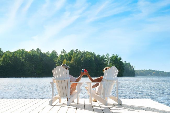 Twee mensen klinken hun glazen op witte stoelen aan een zonnig meer met een beboste oever.