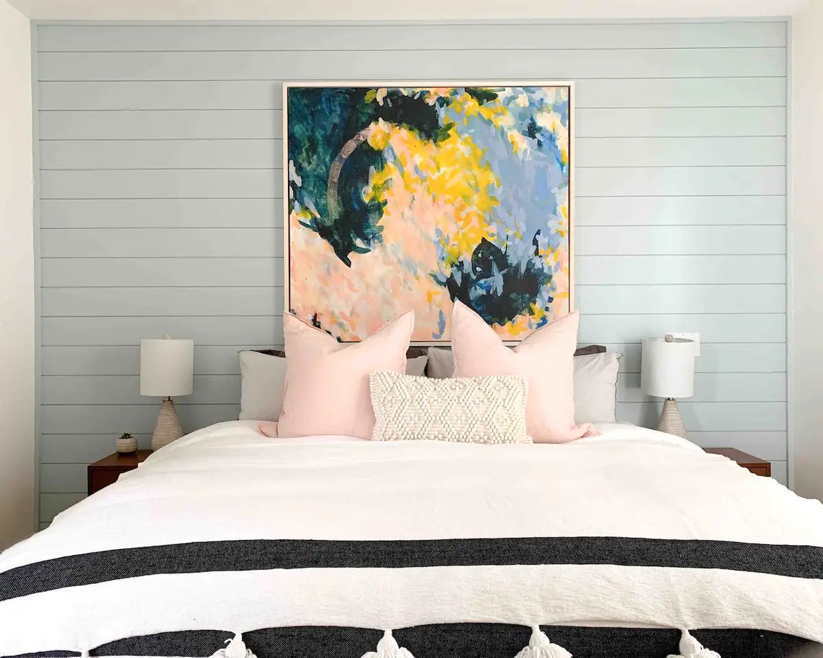 Moderne slaapkamer met wit beddengoed, roze kussens, nachtlampjes en abstracte, kleurrijke kunst boven het bed.