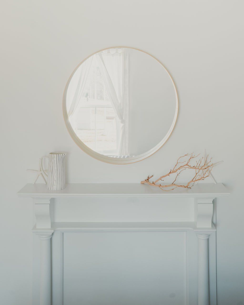 Minimalistische witte schouw met ronde spiegel, klein vaasje en gedroogde takken in een lichte kamer.