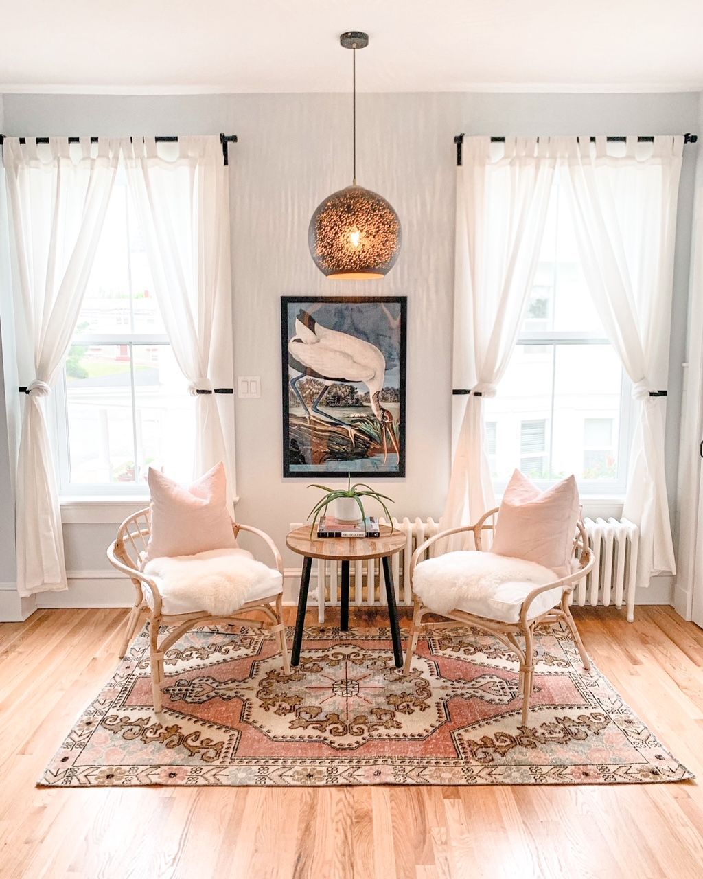 Lichte, luchtige woonkamer met twee witte stoelen, een ronde tafel, een vloerkleed en ingelijste kunst aan de muur tussen twee ramen.
