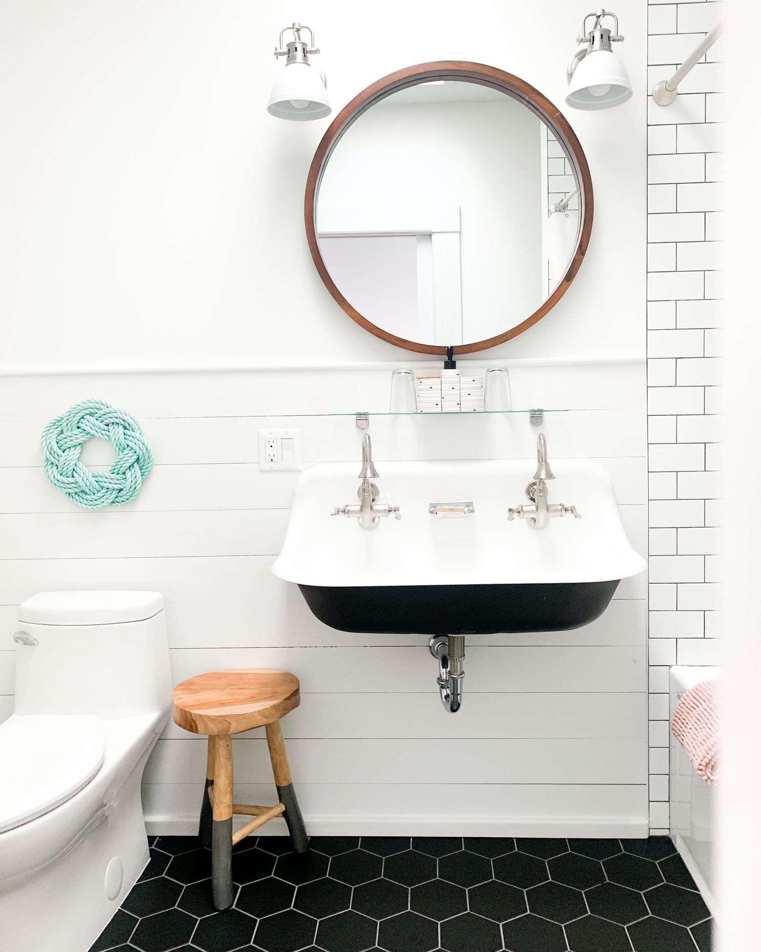 Lichte badkamer met ronde spiegel, witte wastafel, toilet, toiletpot en zwarte zeshoekige vloertegels.