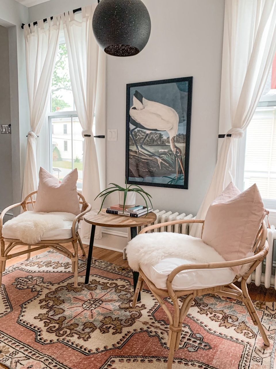 Gezellige zithoek met veel zonlicht, twee rieten stoelen, roze kussens, een vloerkleed met patroon en ingelijste kunstwerken aan de muur.