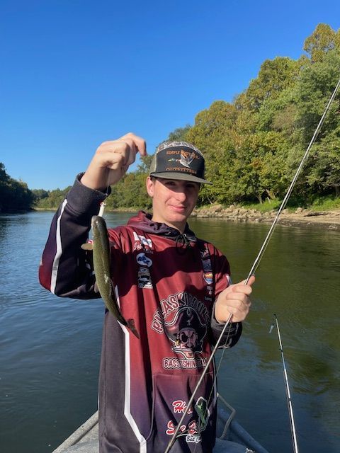 Person in a boat holds up a small fish. Fishing rod visible. Outdoors, river setting with trees.