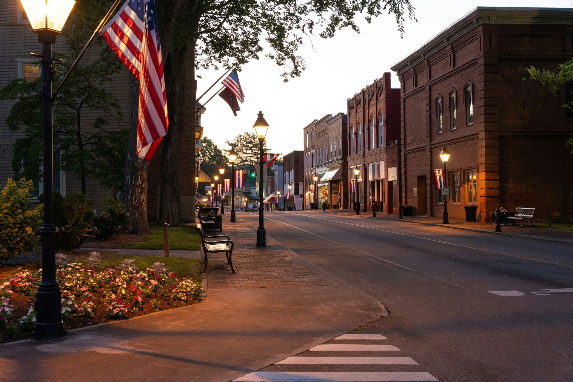 Small-town main street at dusk with American flags, brick storefronts, and a bench along the sidewalk.