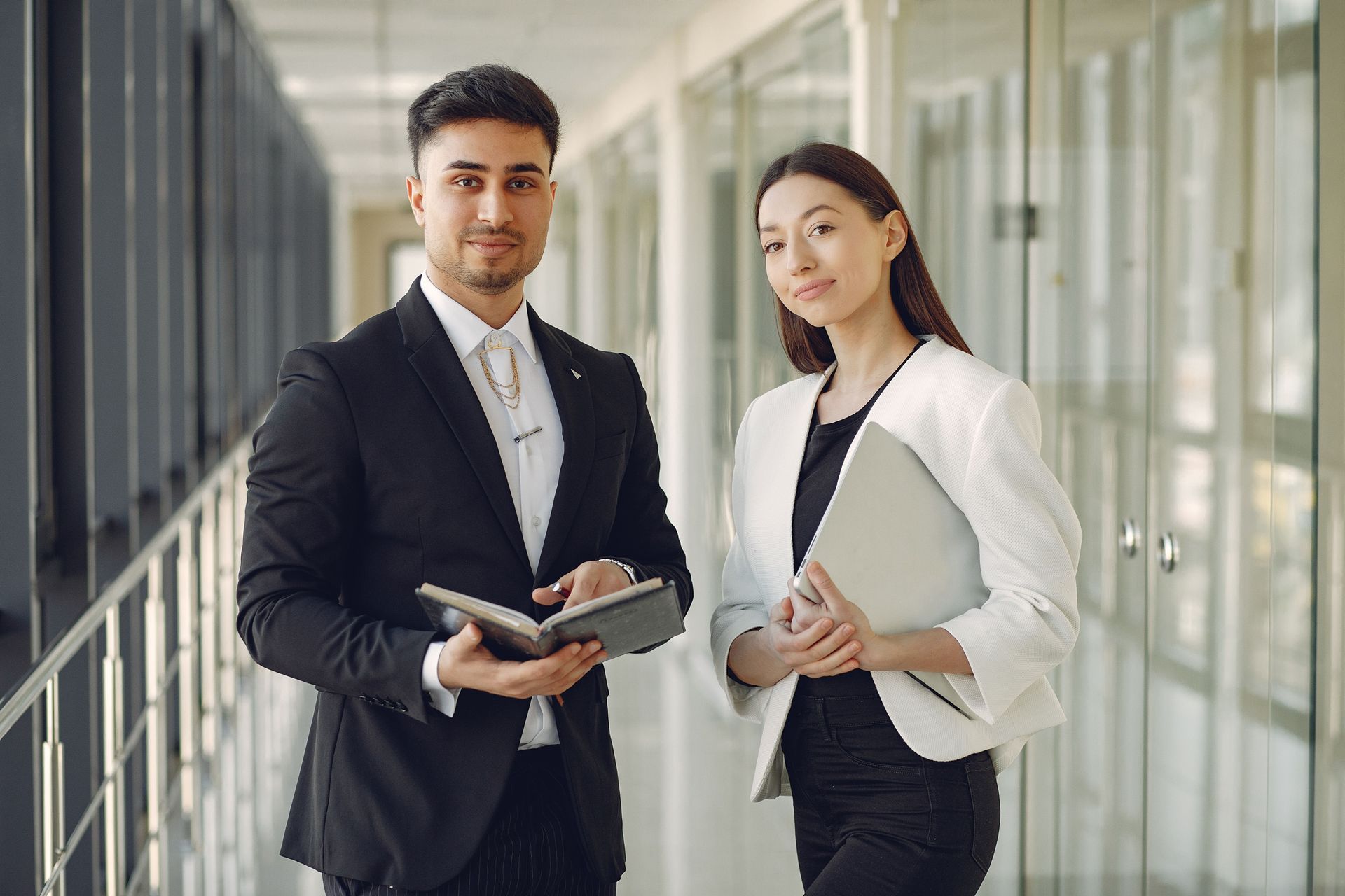 Two professionals in a modern office corridor, one holding a tablet and the other a notebook.