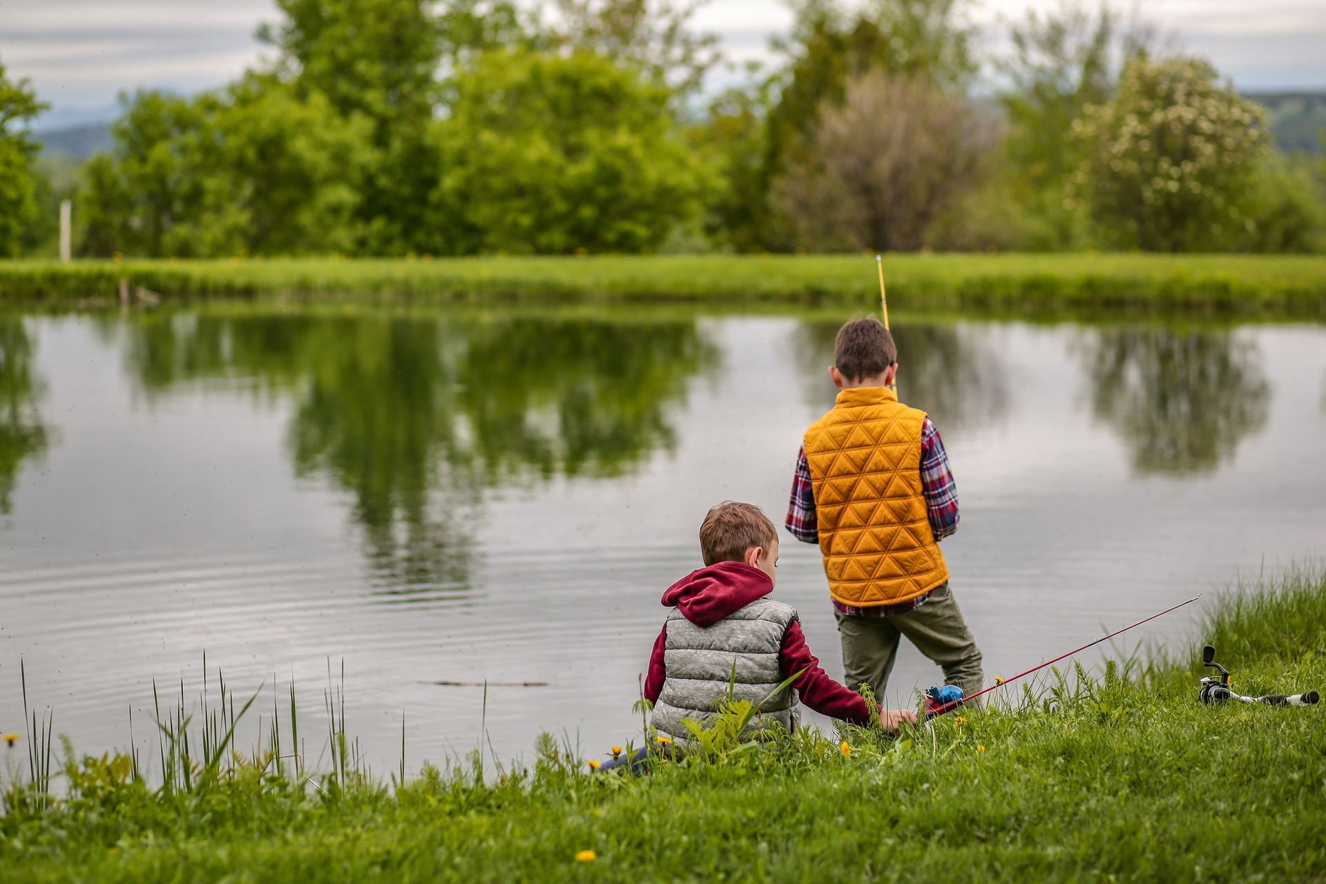 Two children by a calm lake, one standing in an orange vest and one seated in a red jacket.
