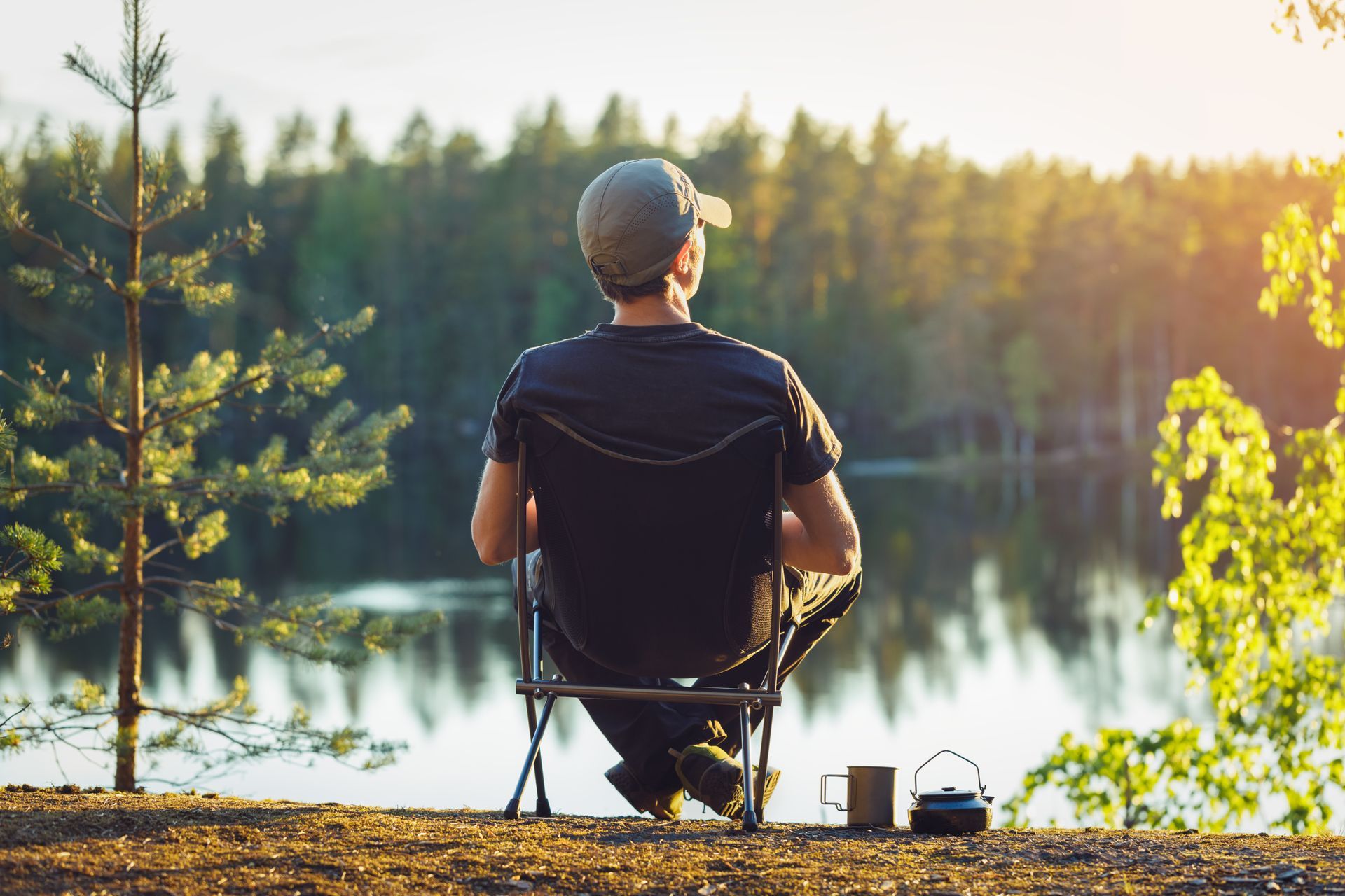 Person fishing from a lakeside chair at sunrise, with rods beside a calm forest lake.
