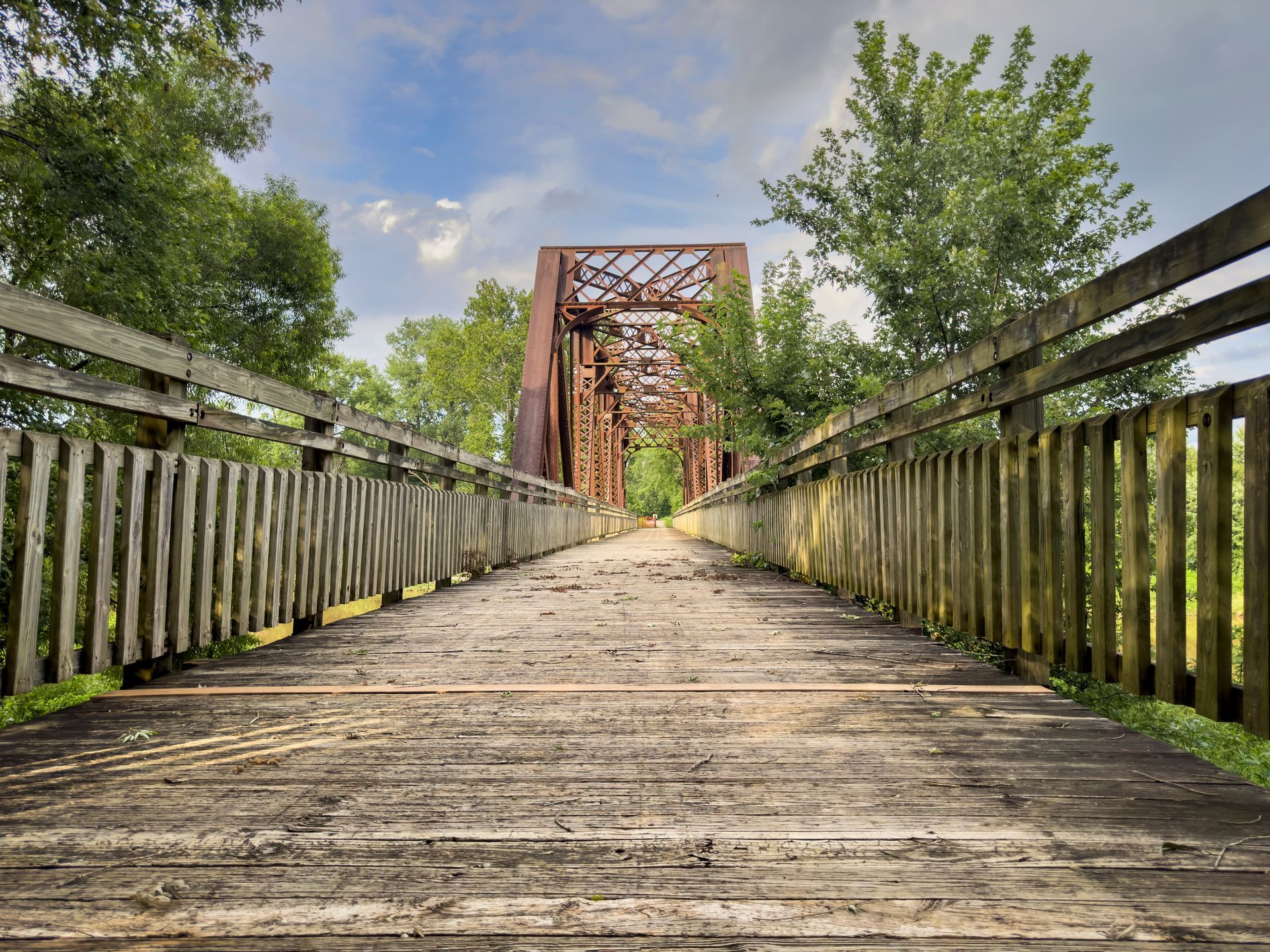 Wooden bridge walkway leading toward a rusty truss bridge under a partly cloudy sky, lined with green trees.