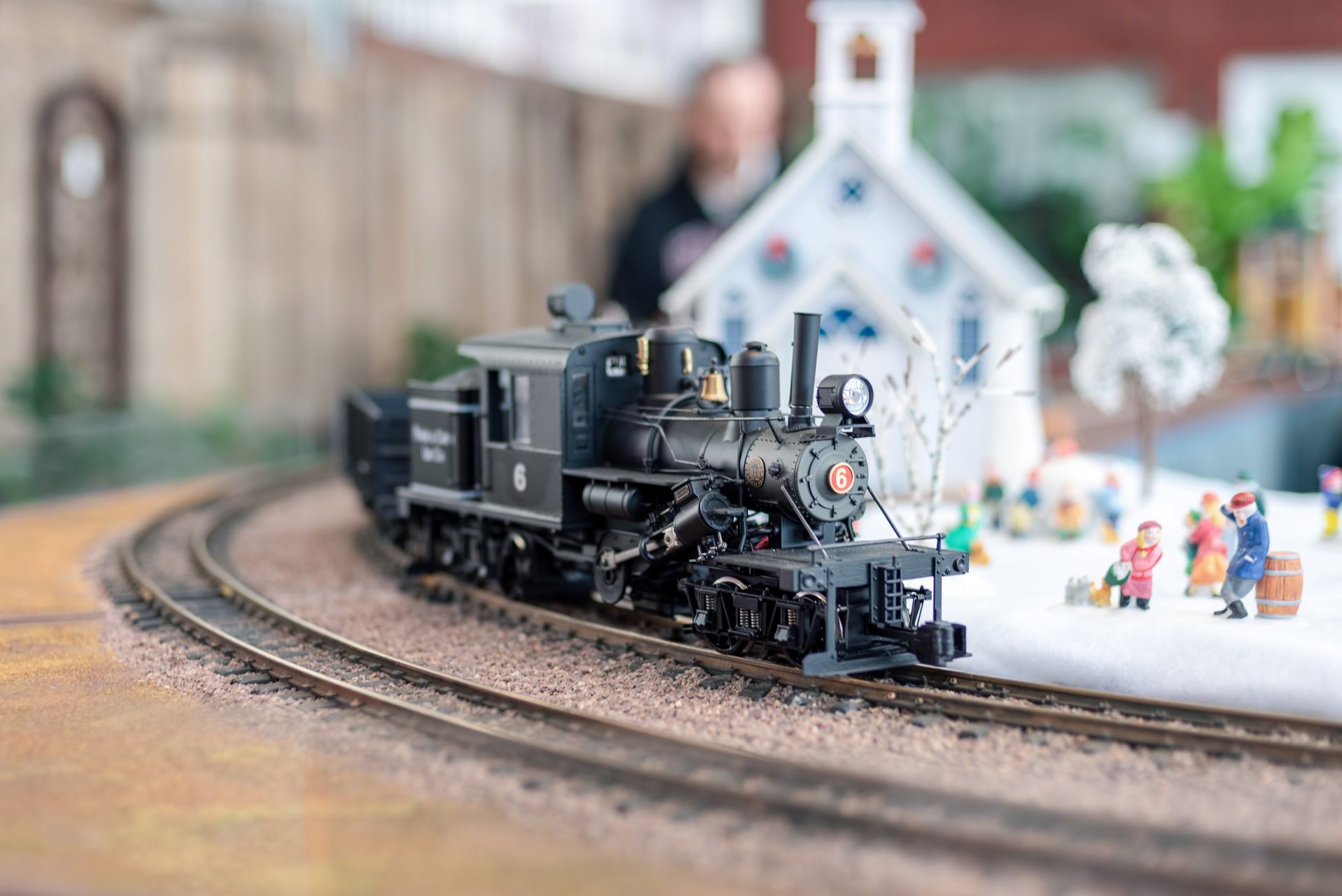 Black model steam locomotive on curved track beside a small white church with miniature figures nearby