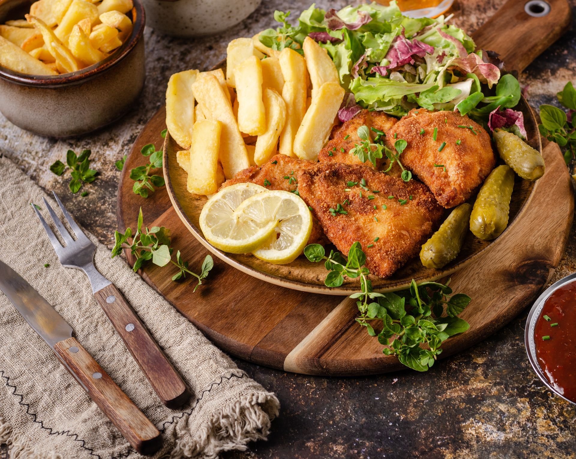 Fried chicken with fries, lemon and salad on a wooden board, with dipping sauce and cutlery nearby