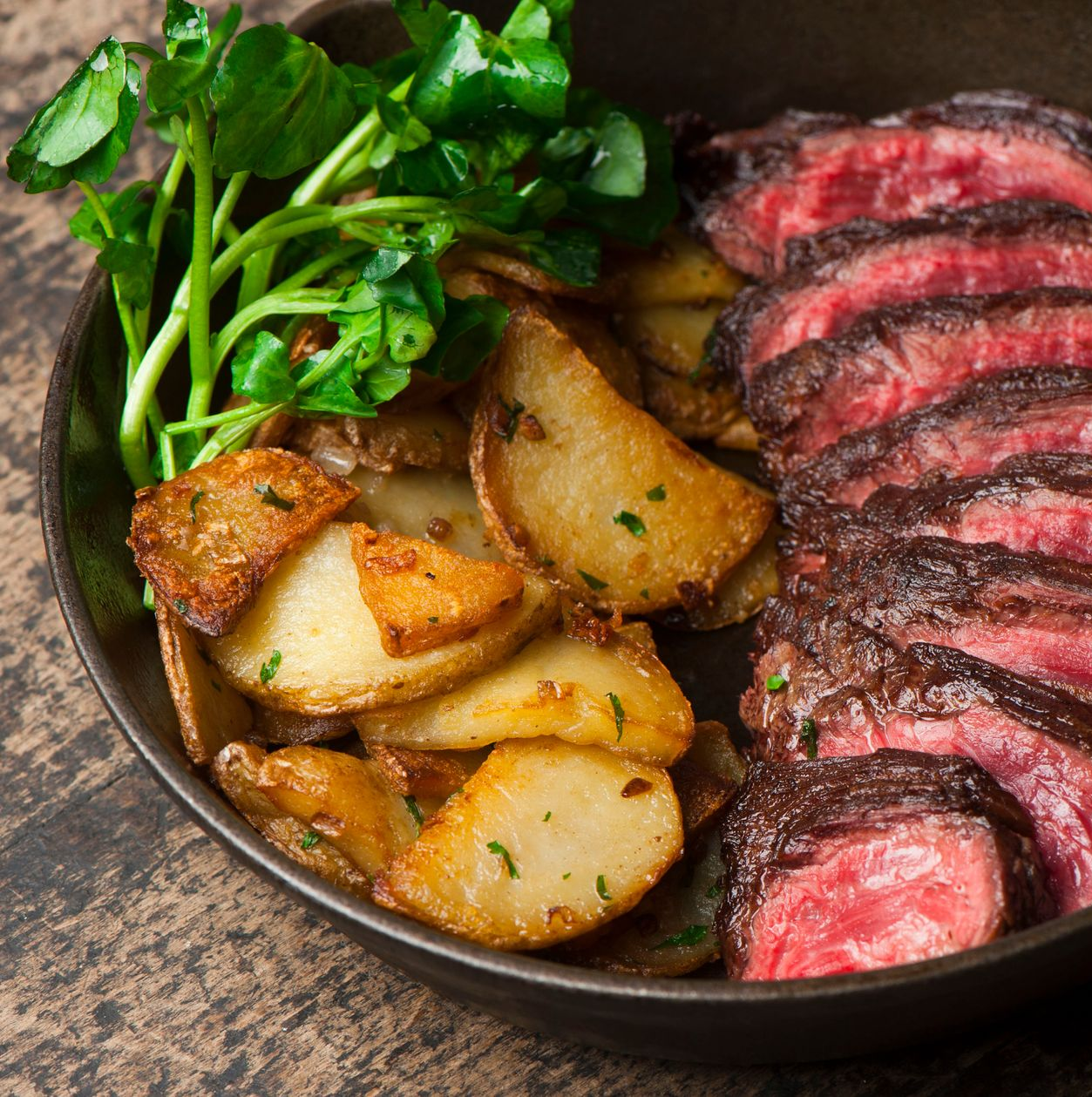 Seared steak with roasted potatoes and greens in a black bowl on a rustic table