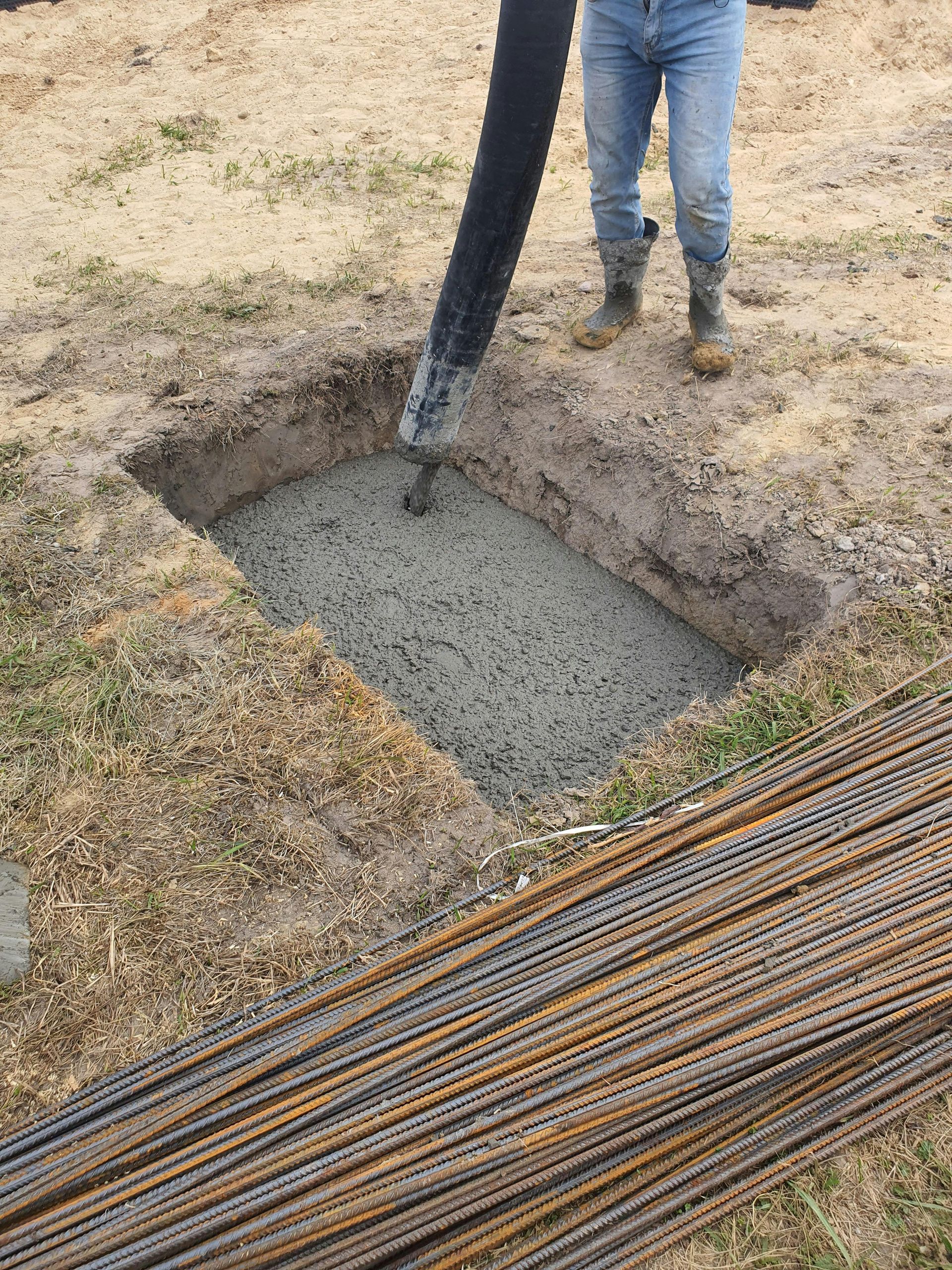 Concrete being poured into a square hole beside a bundle of rebar on a dirt construction site