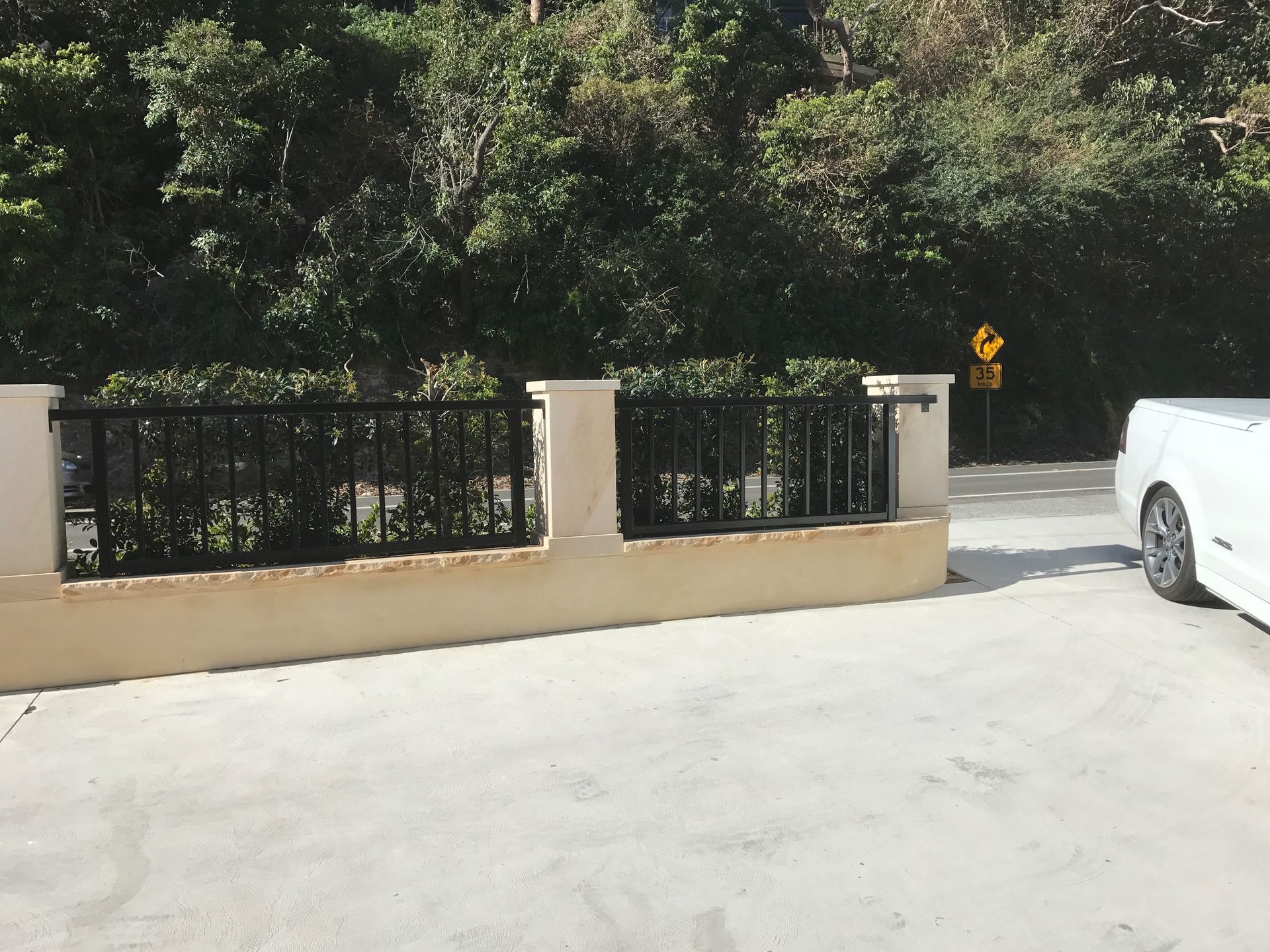 Tan wall with black railing, greenery behind it, and a white car parked on a concrete surface.