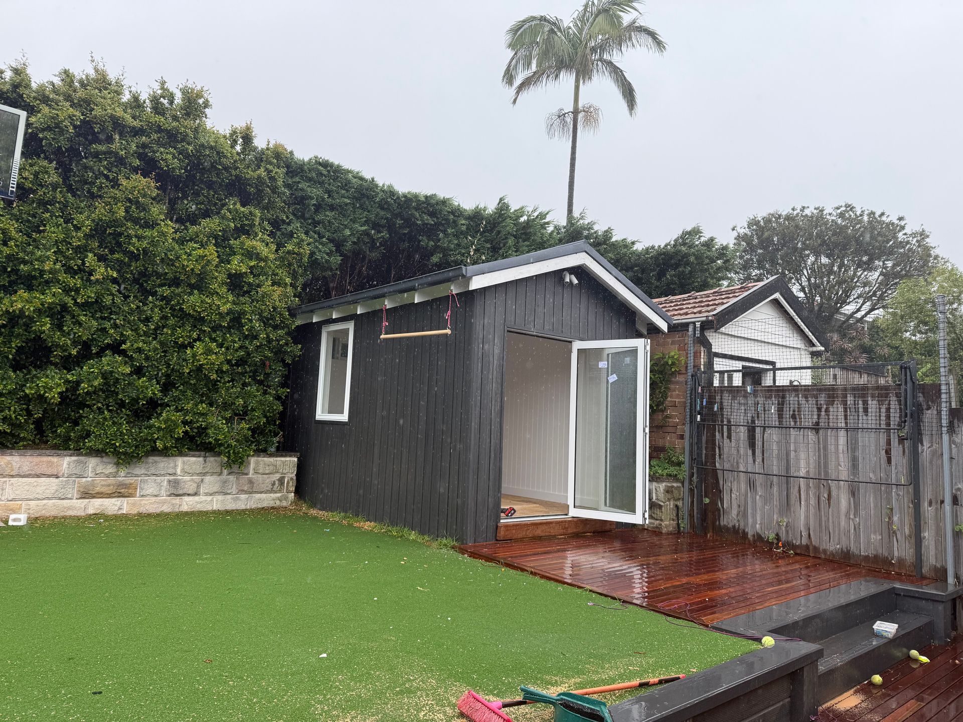Black shed with open white doors on a green lawn with a wooden deck.