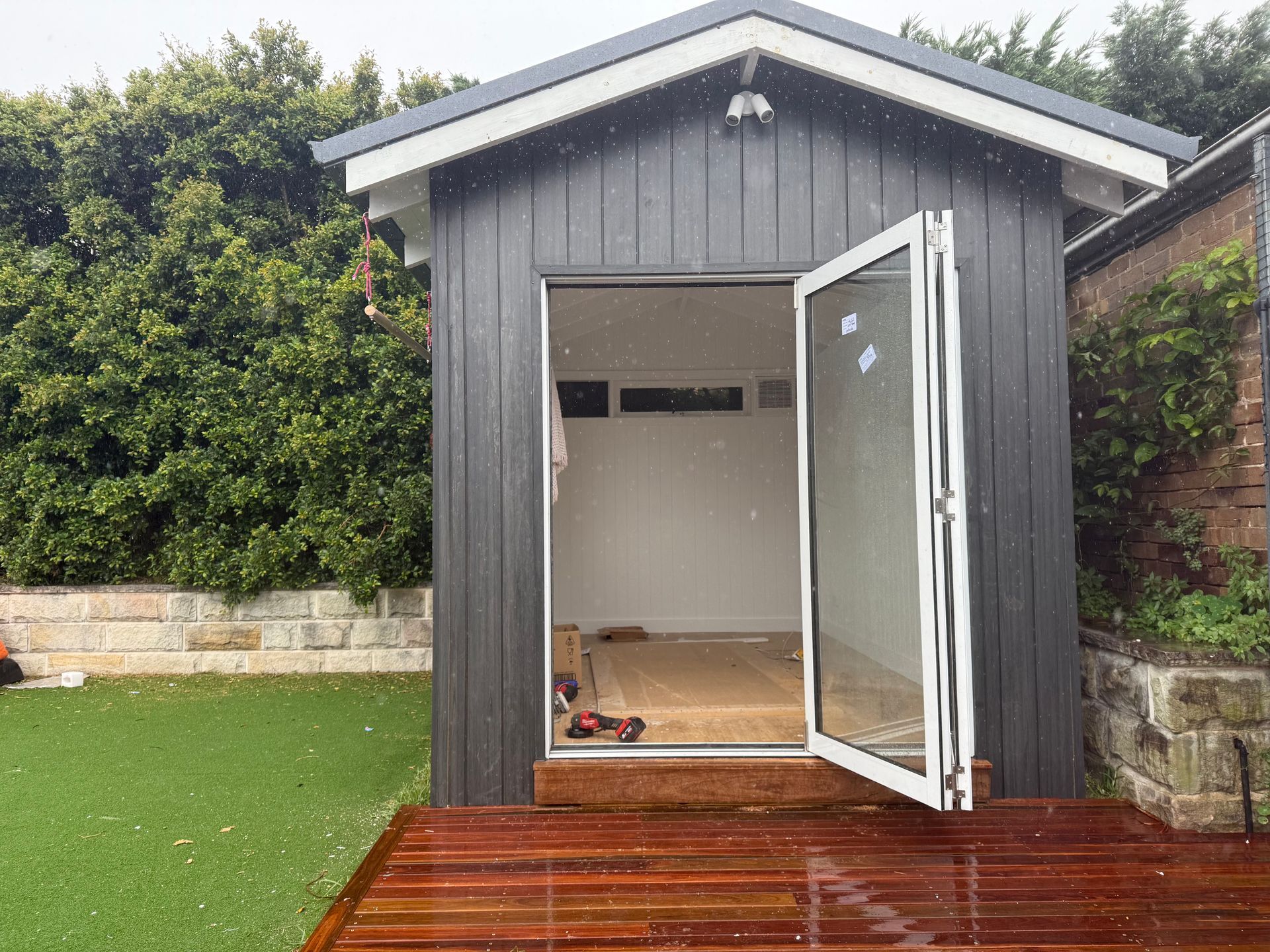 Dark gray shed with open white doors, on a wooden deck. Green grass and stone wall in the background.