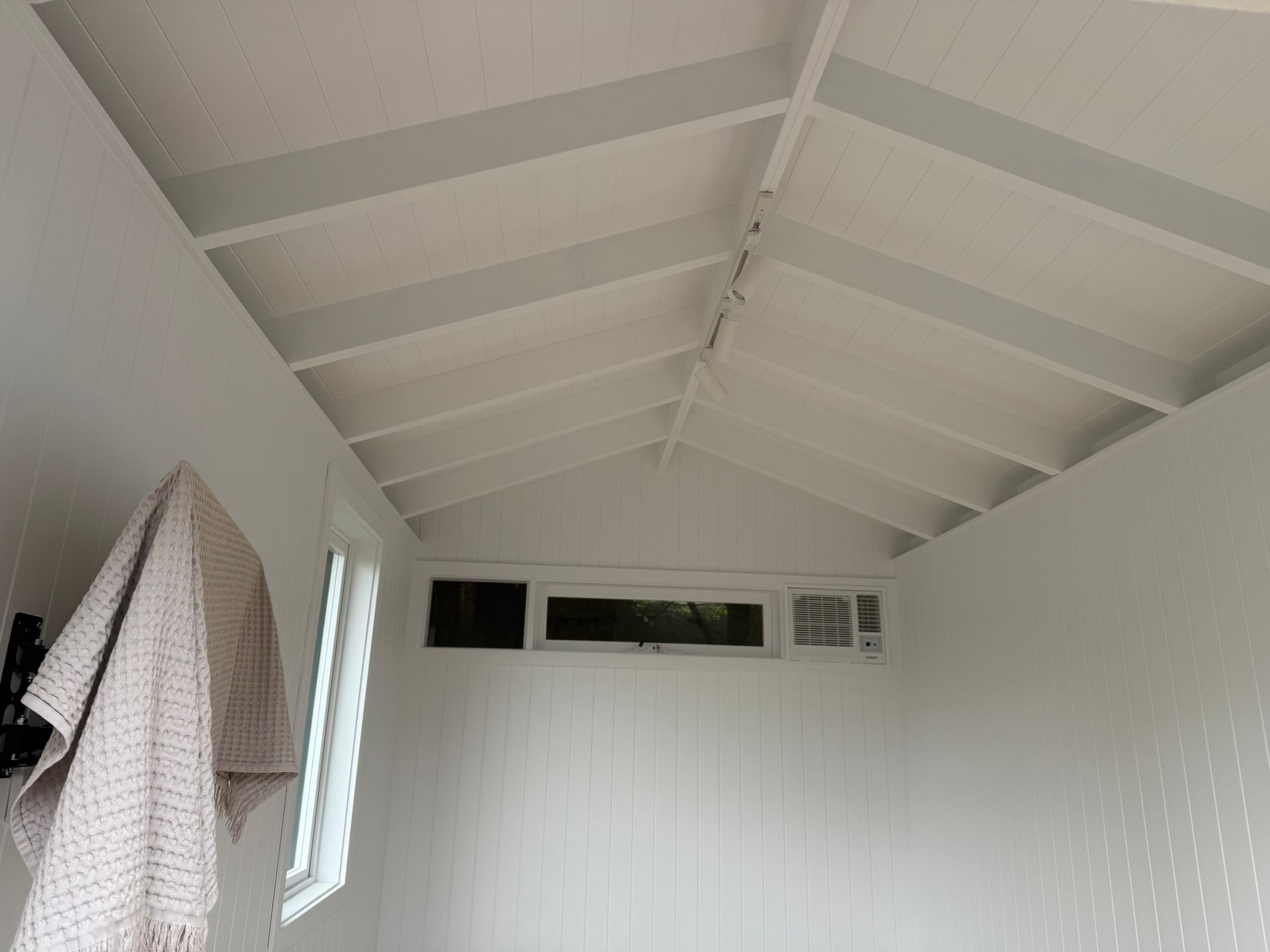 White interior with a sloped, beamed ceiling. A window and a wall-mounted towel are visible.