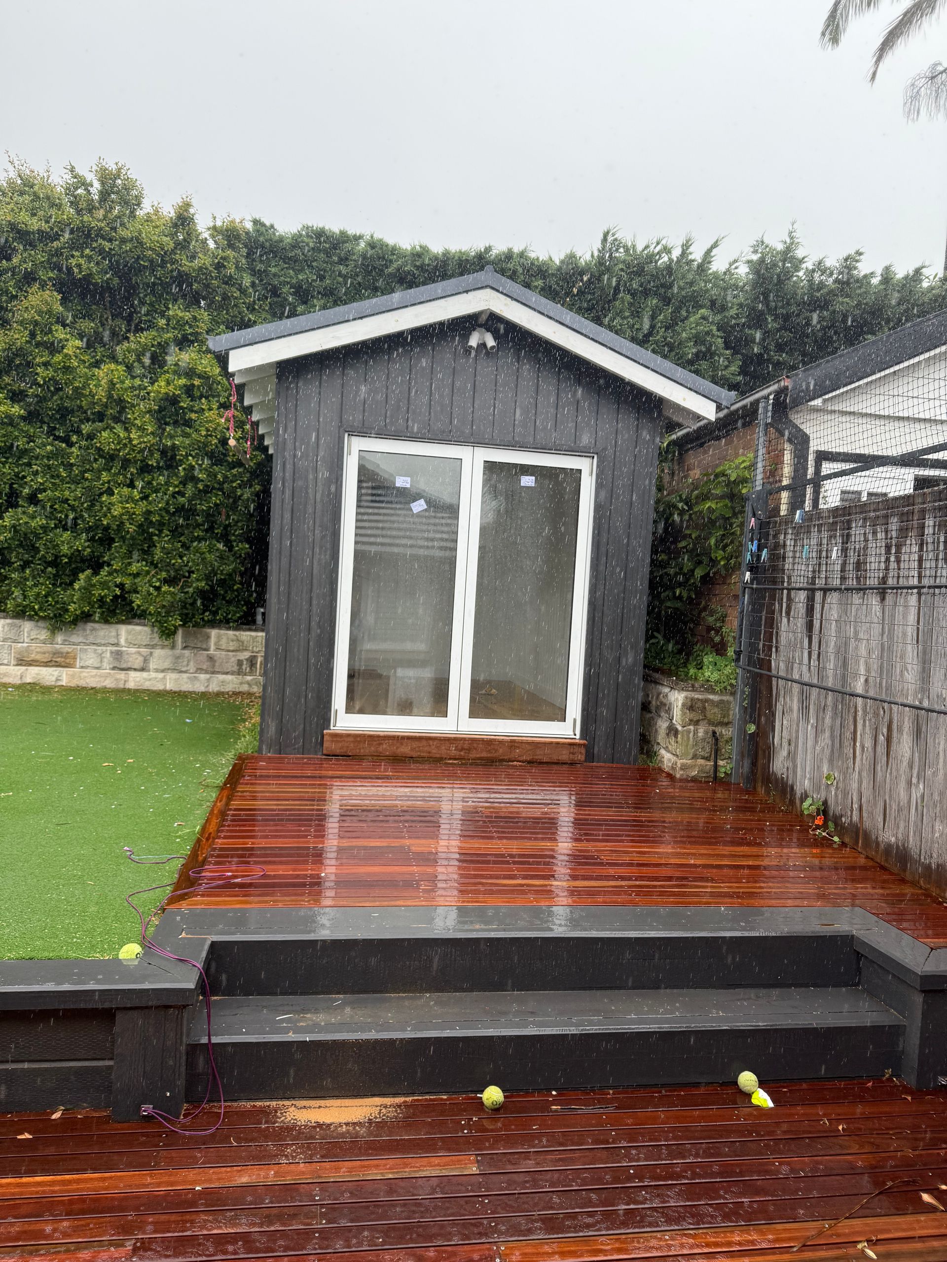 Black shed with white-framed glass doors on a wooden deck. Green lawn and steps lead up to the deck.