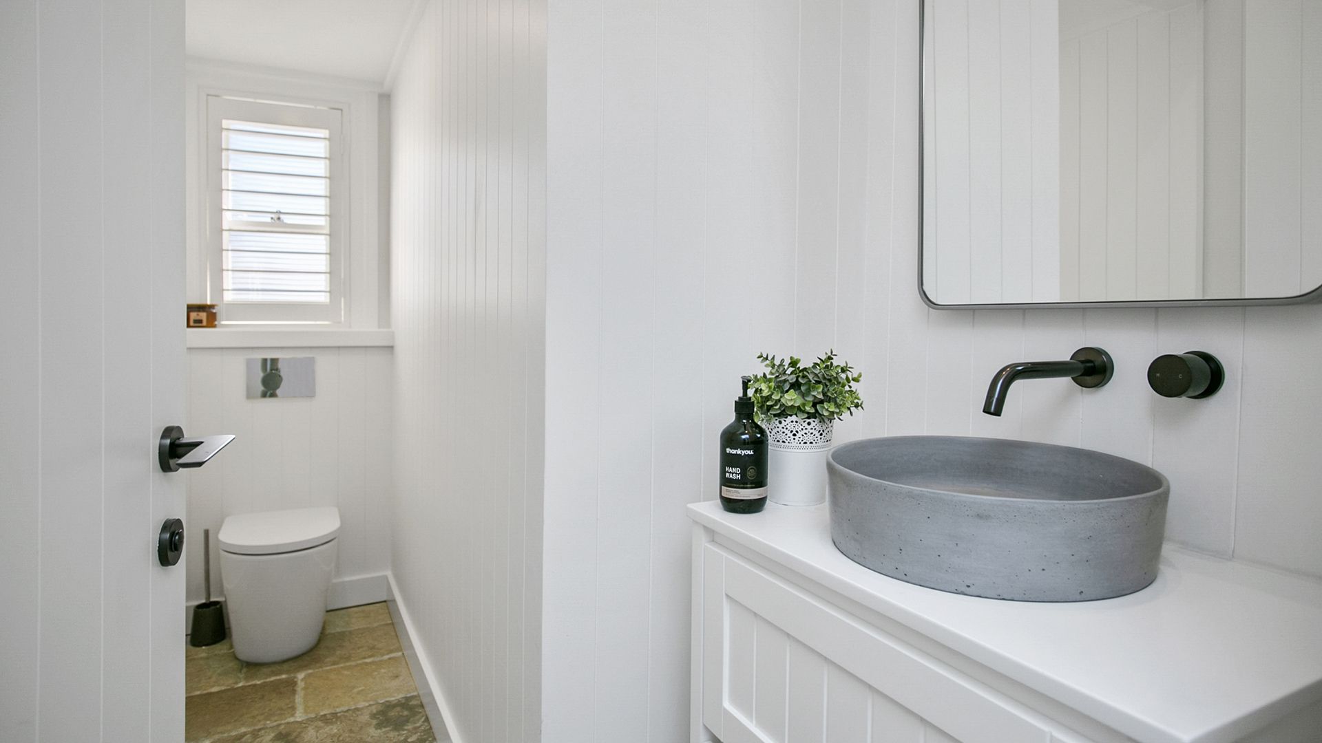 Small, white bathroom with toilet, sink, and shuttered window. Gray stone floor and white walls.