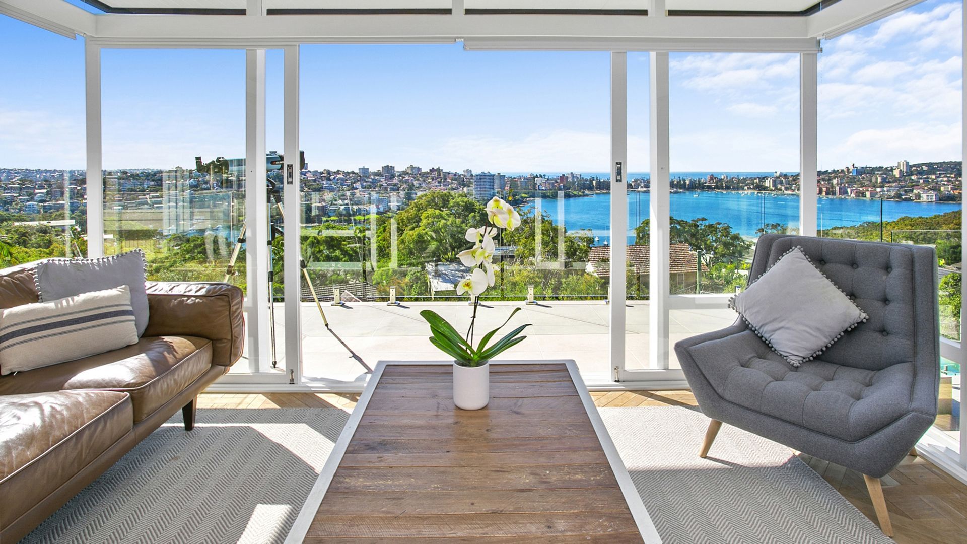 Sunroom with city and water views; leather couch, gray chair, coffee table, white orchid.