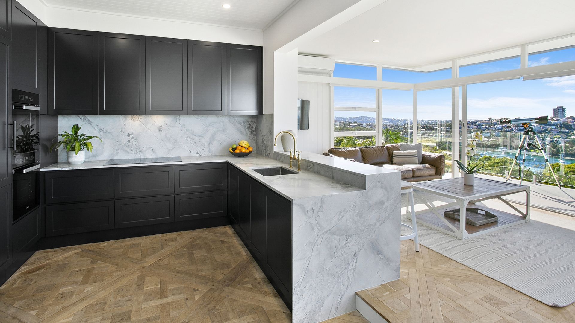 Modern kitchen with black cabinets, marble countertops, and city view through large windows.