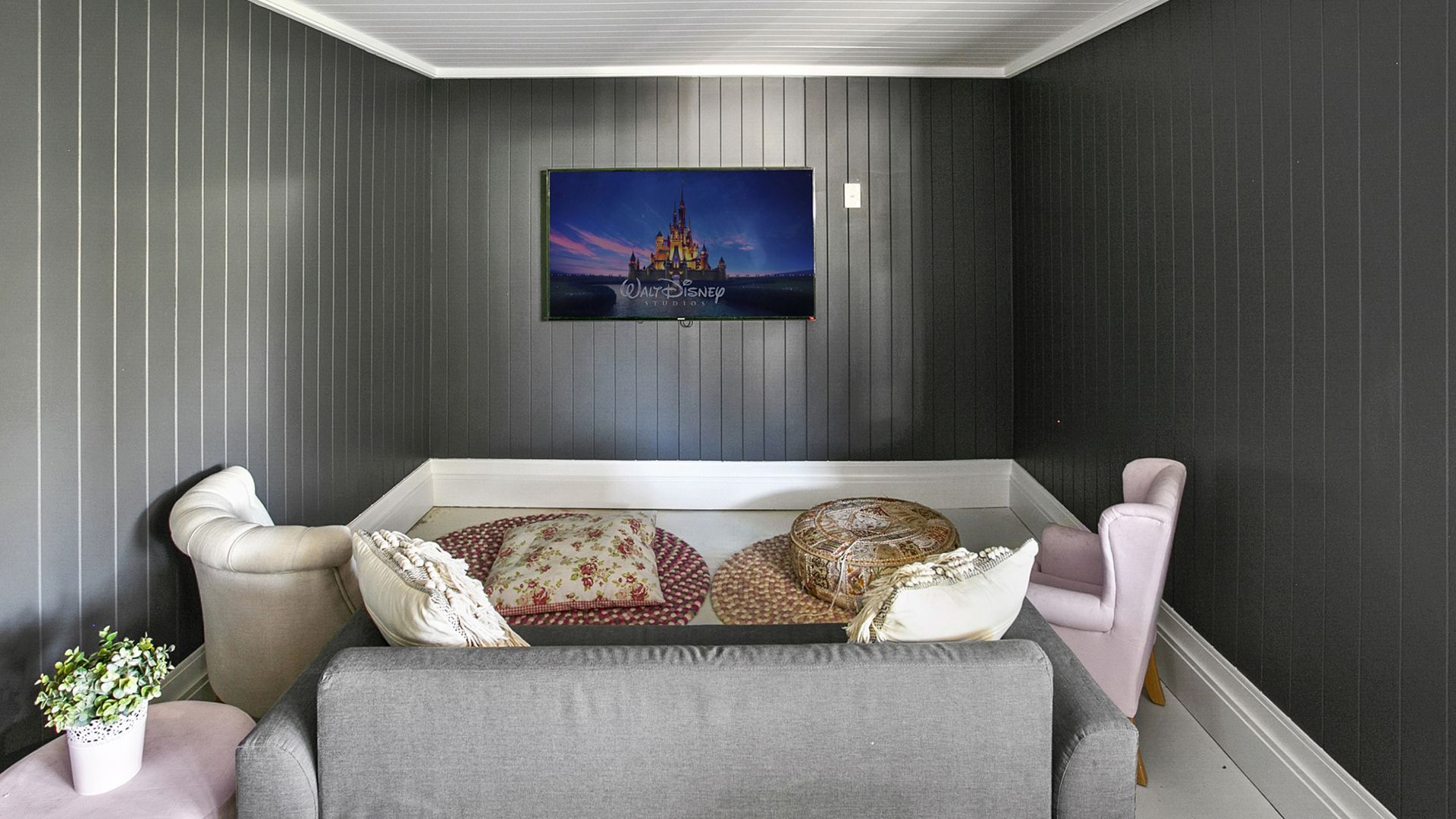 Cozy gray room with a TV above a gray couch, flanked by two pink chairs, and paneled walls.