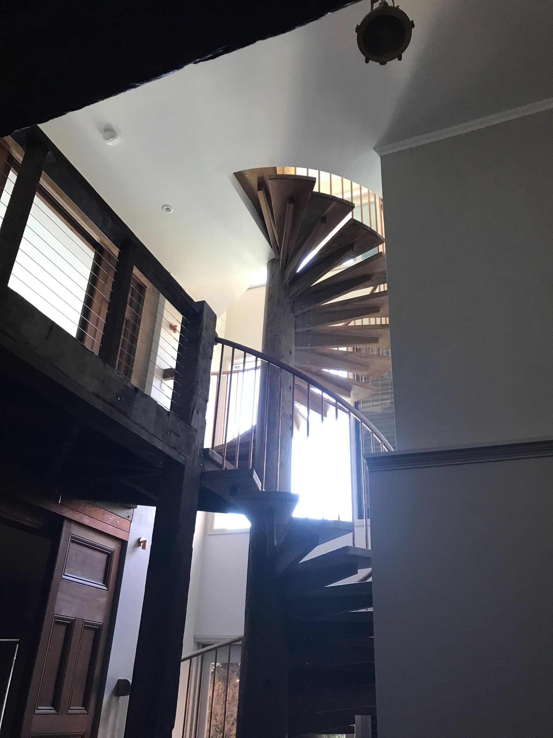 Wooden spiral staircase ascending to a white ceiling, viewed from below. Dark railing.