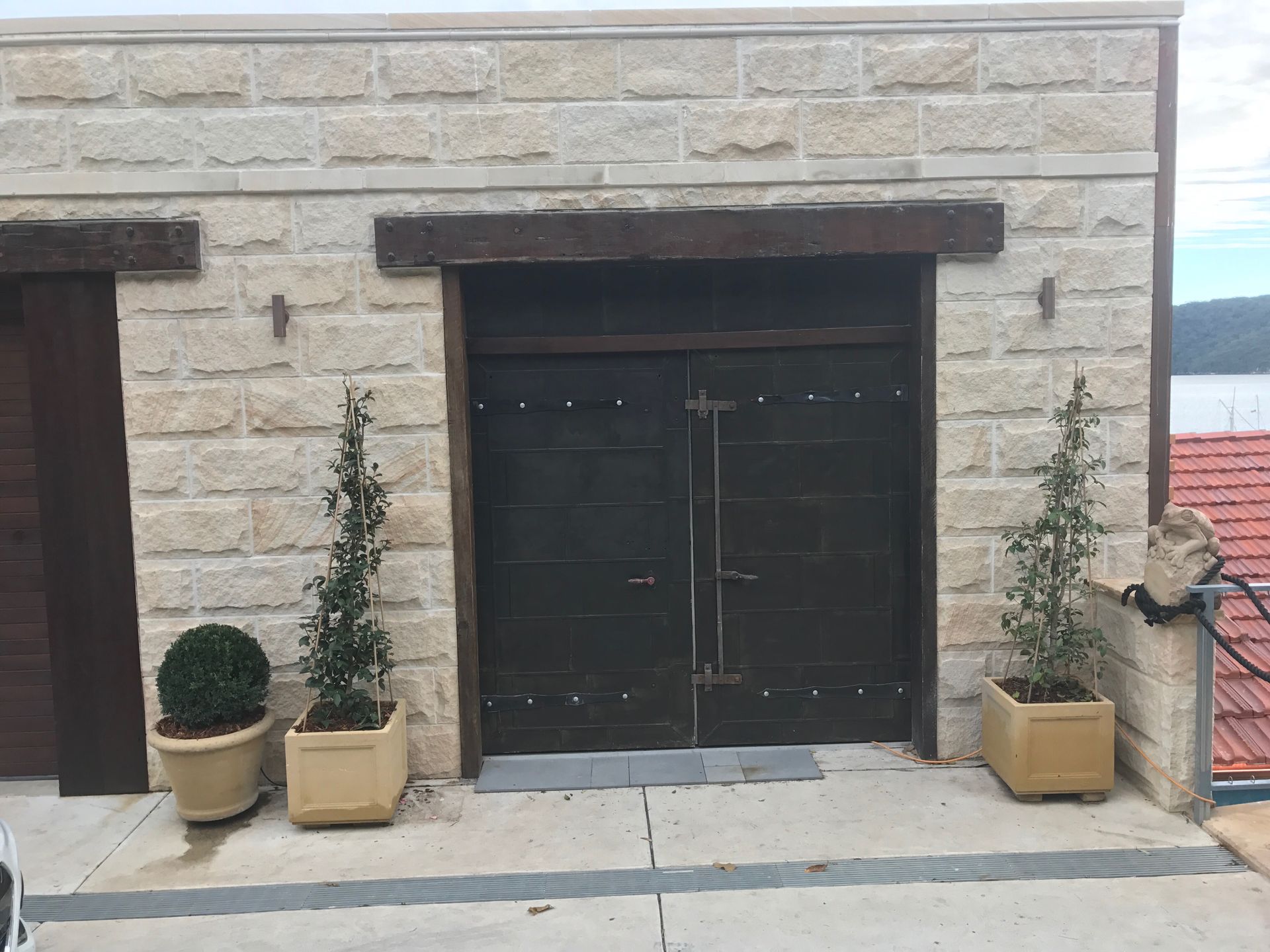 Tan stone building with dark brown doors, topped by a wooden beam. Potted plants flank the doorway.