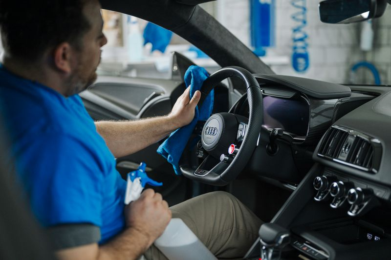 Mechanic cleaning a car dashboard and steering wheel with a blue cloth inside a vehicle