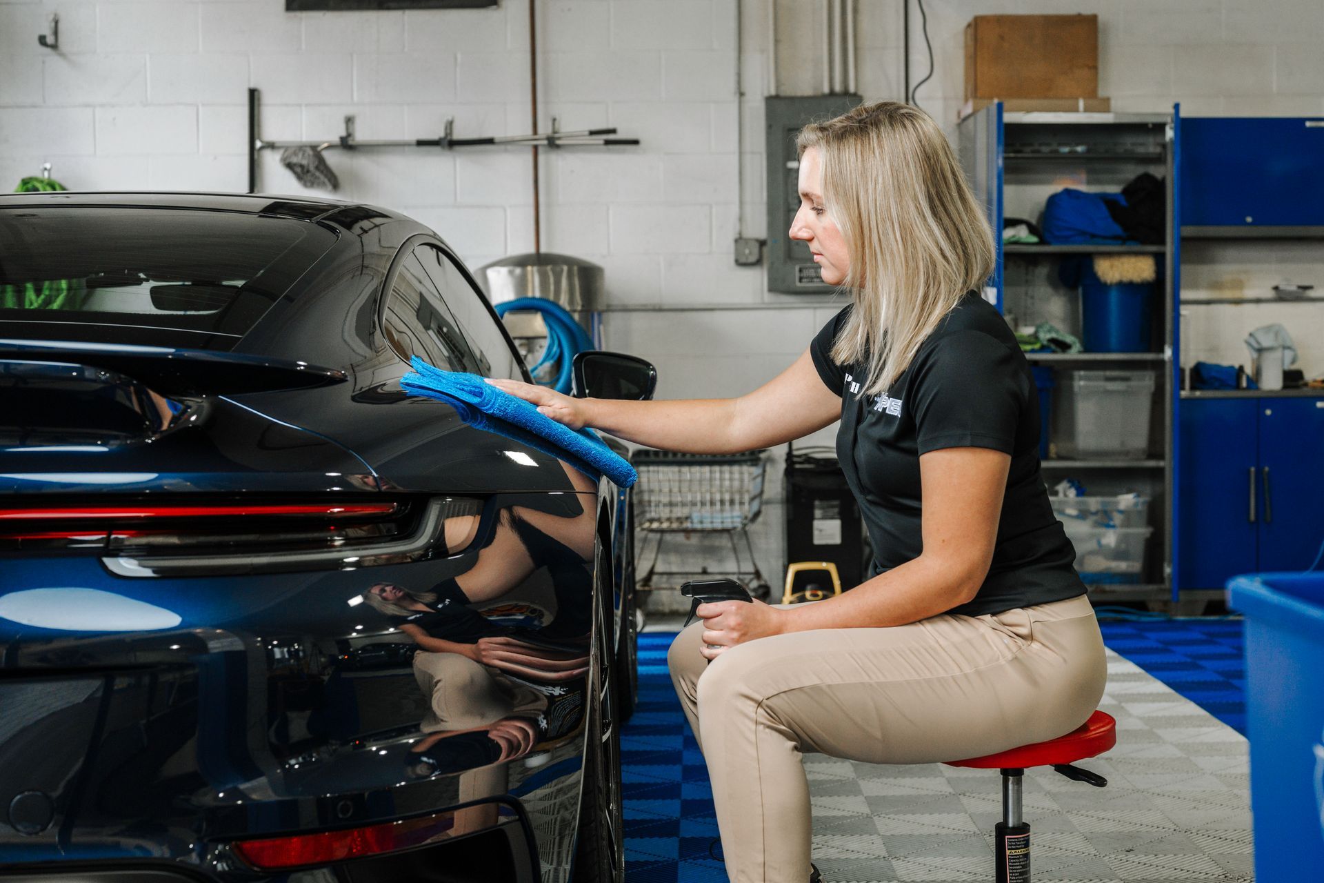 Woman polishing a black car in an auto repair shop with blue tools and shelves nearby