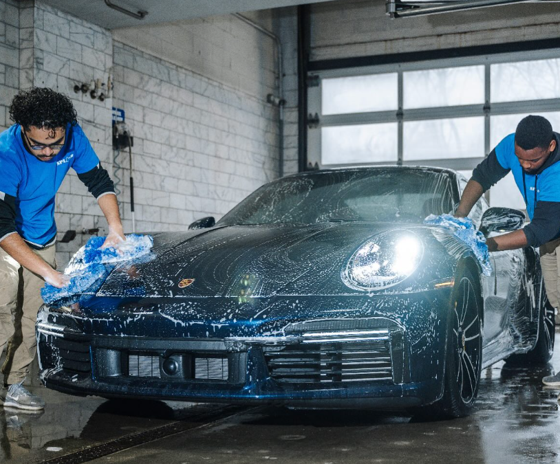 Two people washing a dark blue Porsche with soap and blue cloths inside a car wash bay.