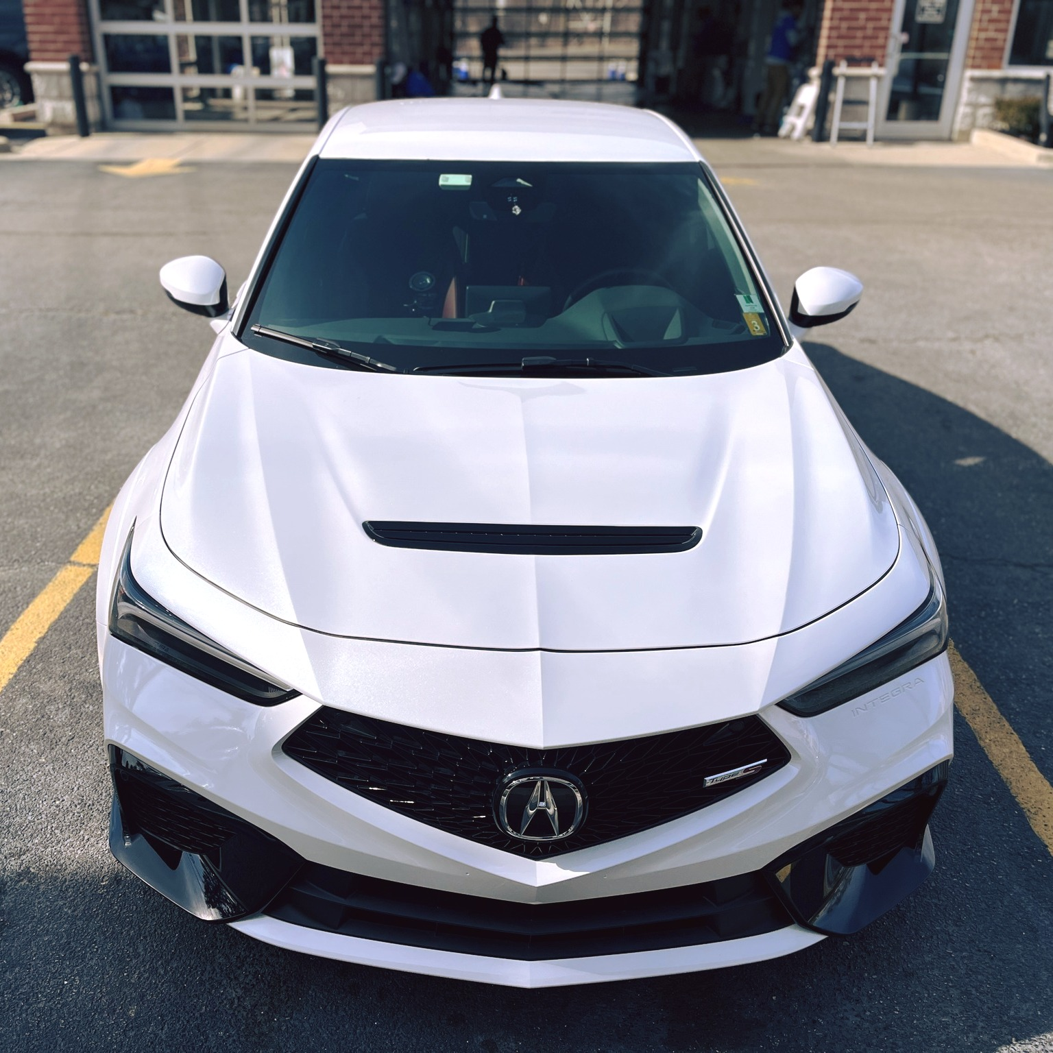 White Acura Integra parked, front view. Black accents, parked in a lot in front of a building with garage doors.