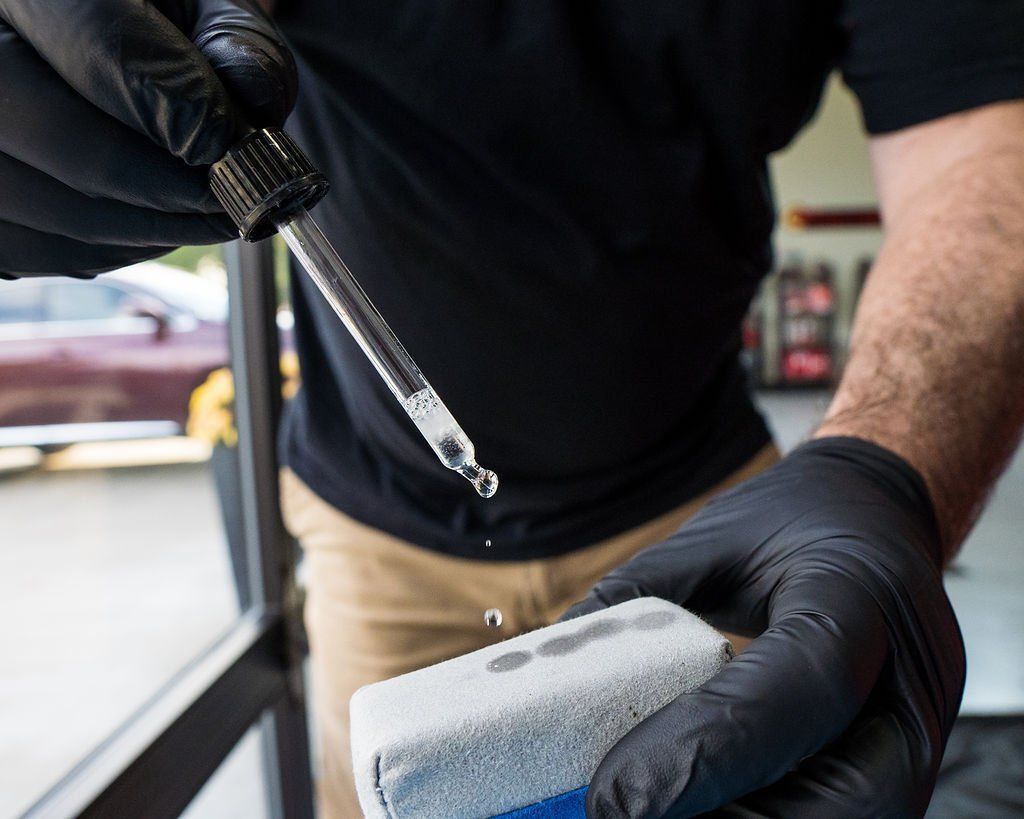 A gloved person dispensing liquid from a dropper onto a white applicator sponge in front of a car.