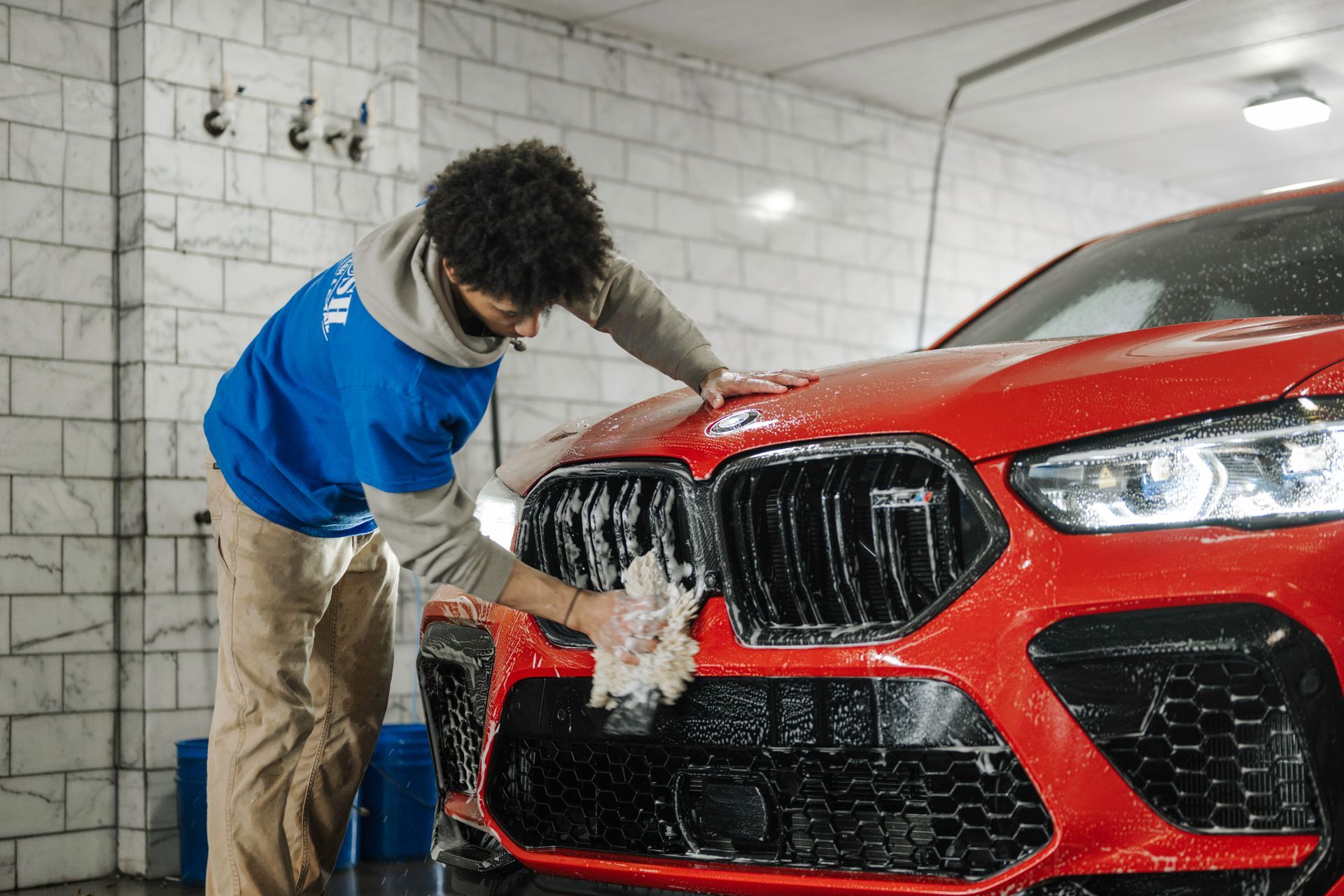 Person washing a red car’s front grille in a tiled auto detail bay