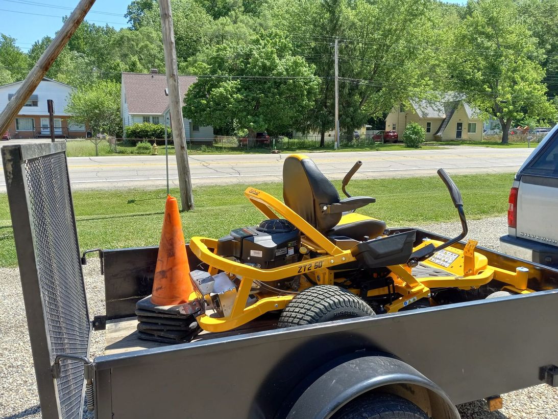 Yellow riding lawn mower on a trailer with an orange cone outdoors.