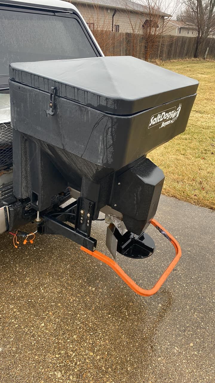 Black snowblower attachment mounted on a vehicle beside a wet driveway, with orange power cord on the ground