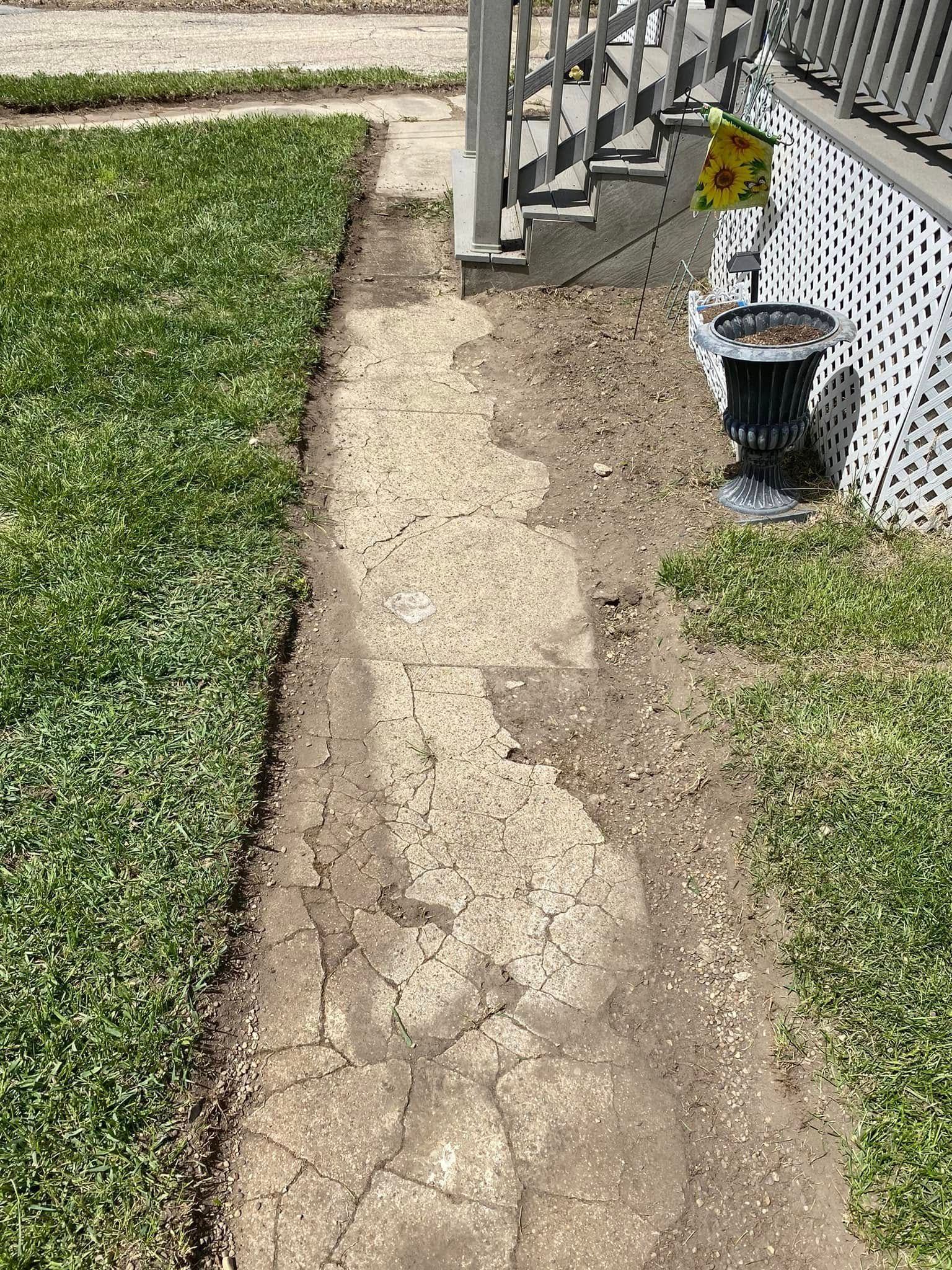 Cracked concrete walkway beside a grass lawn and house steps, with a small potted plant near the porch.