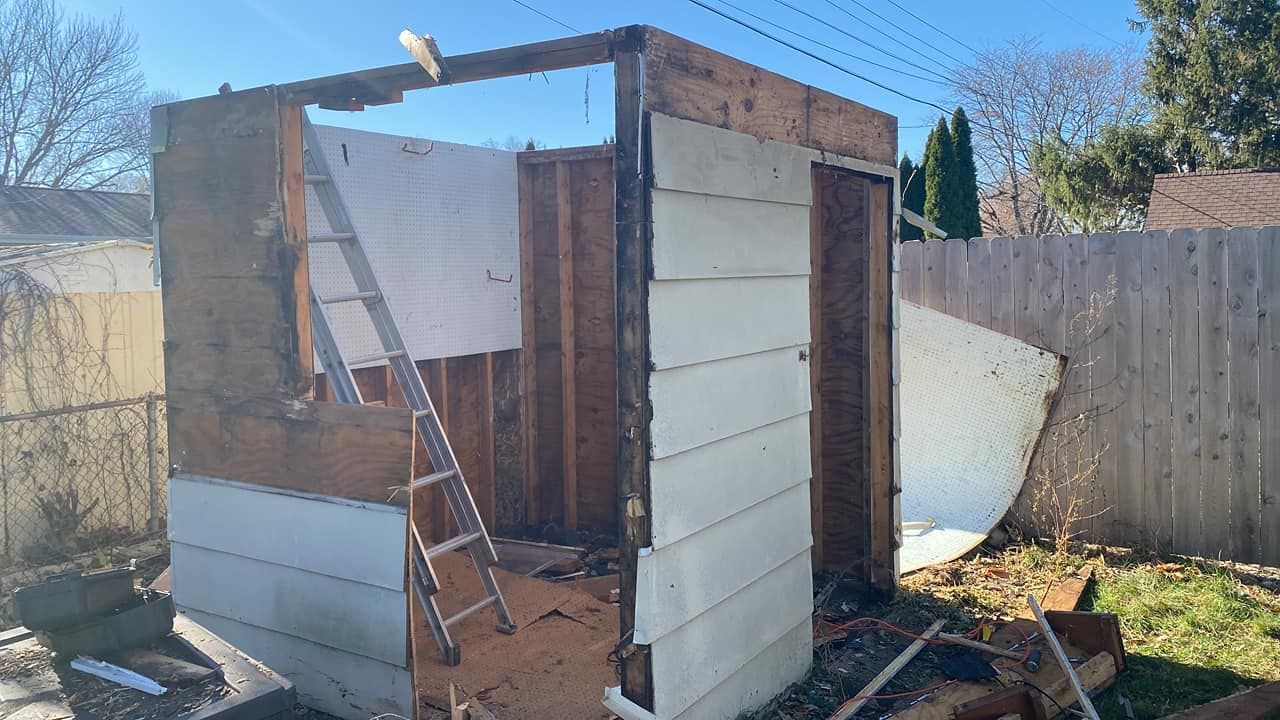 Partially built shed with white siding, open frame, and a ladder in a backyard under construction