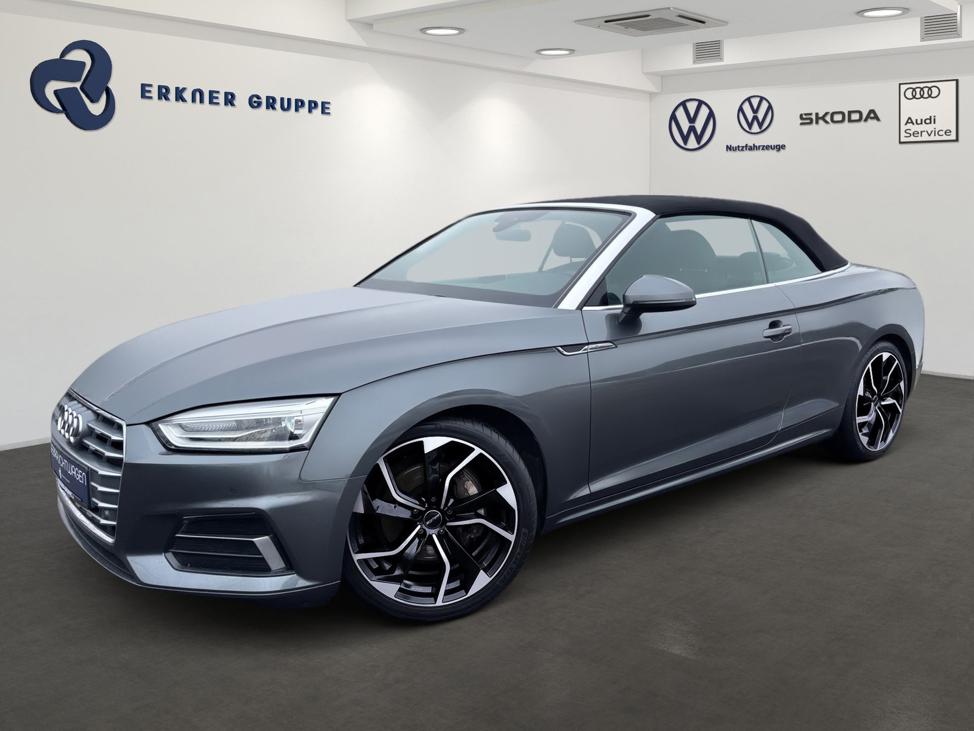 Graues Audi Cabriolet im Ausstellungsraum, schräge Frontansicht, mit schwarzen Felgen und schwarzem Verdeck