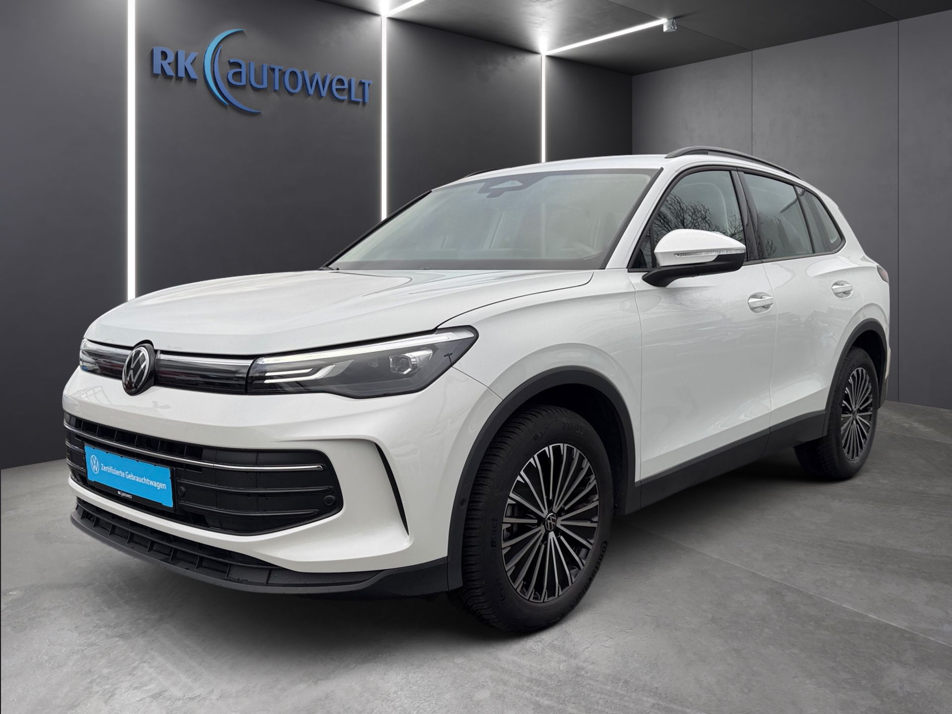 Weißer Volkswagen SUV im Ausstellungsraum, schräge Frontansicht, mit dunklen Felgen und grauer Wand im Hintergrund.