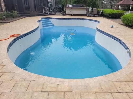 Blue swimming pool in a paved backyard with steps and lounge chairs nearby