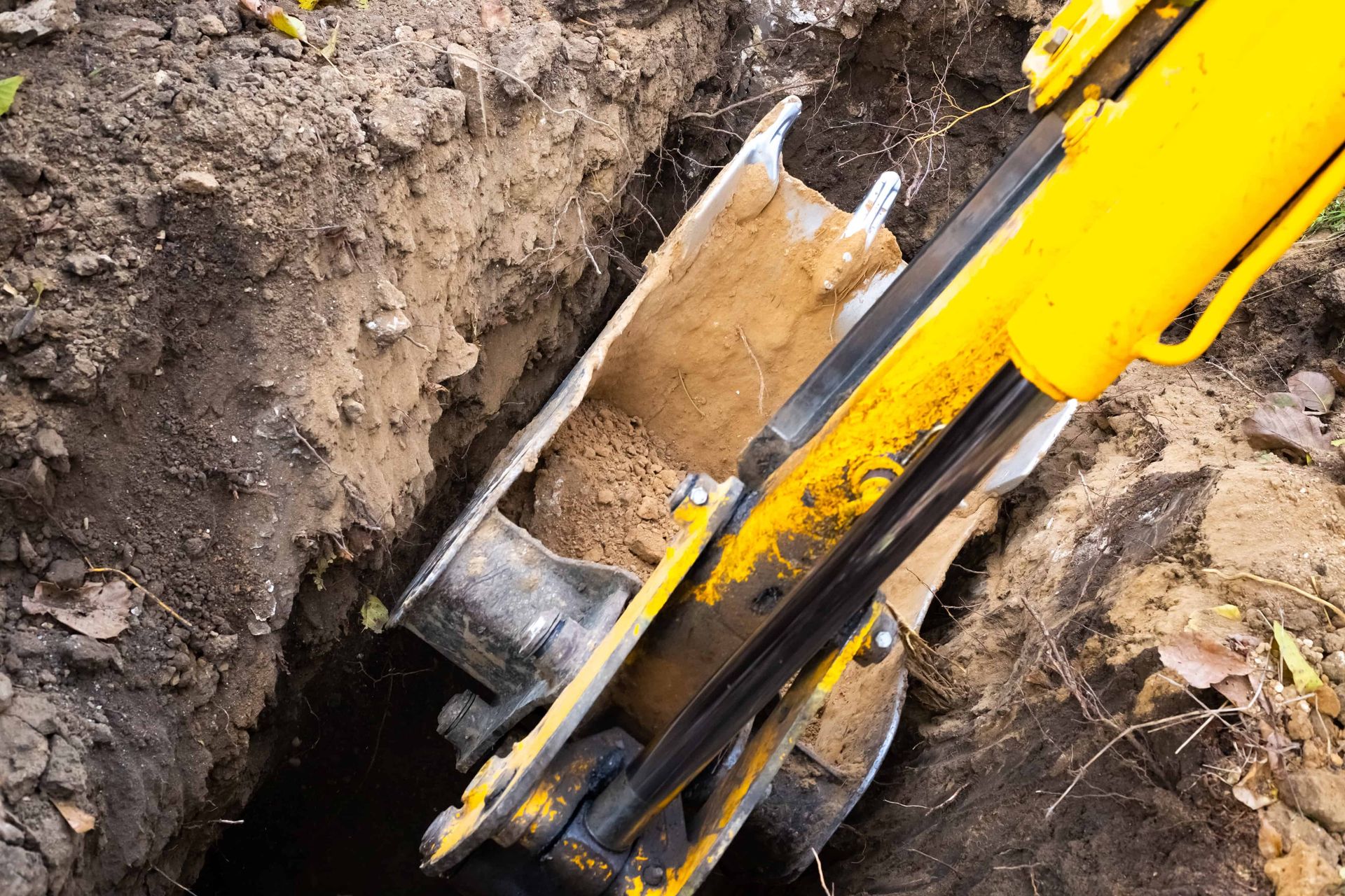 Yellow excavator bucket digging a trench in muddy soil