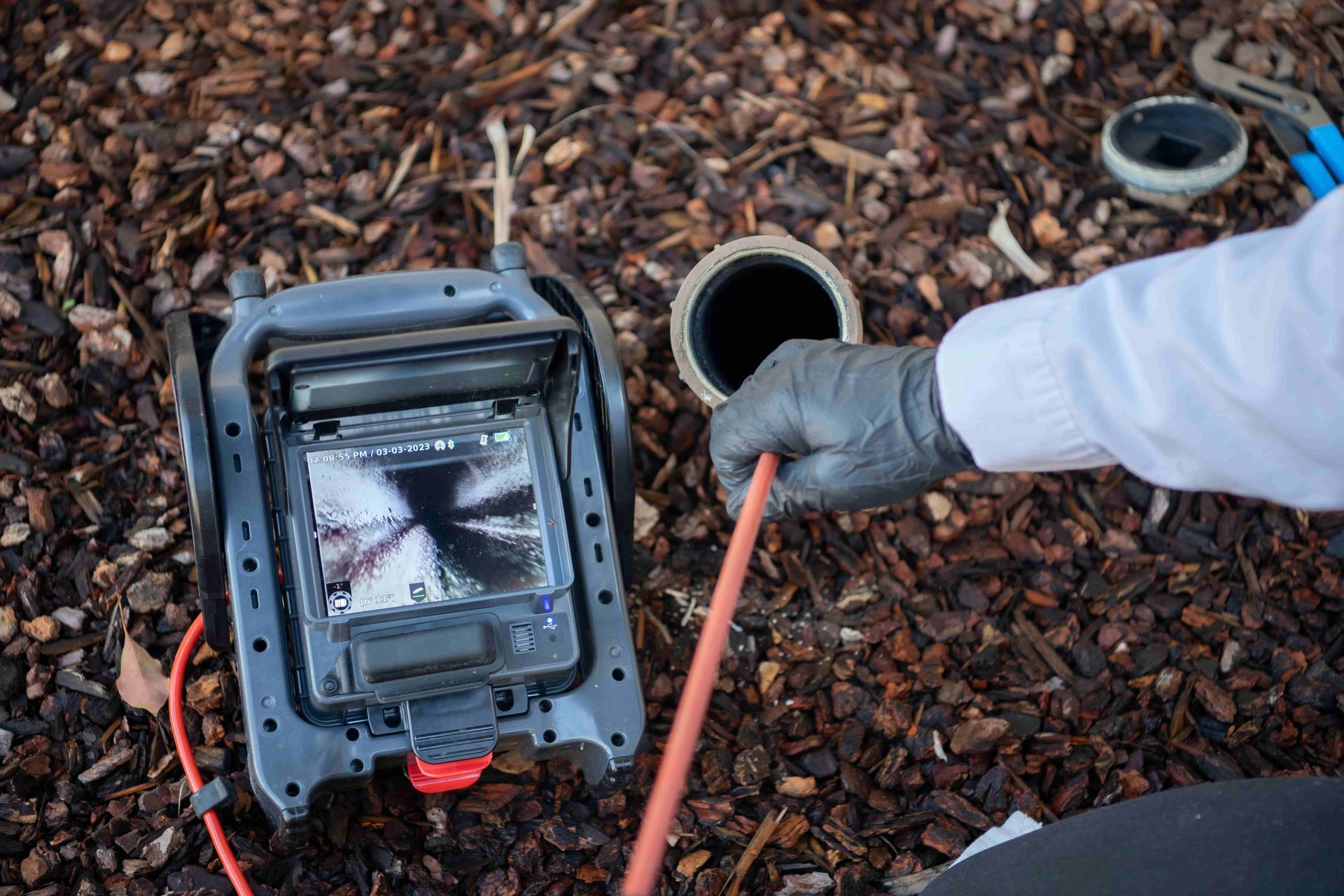 CCTV inspection camera on mulch beside an open pipe, with a gloved hand holding a red cable