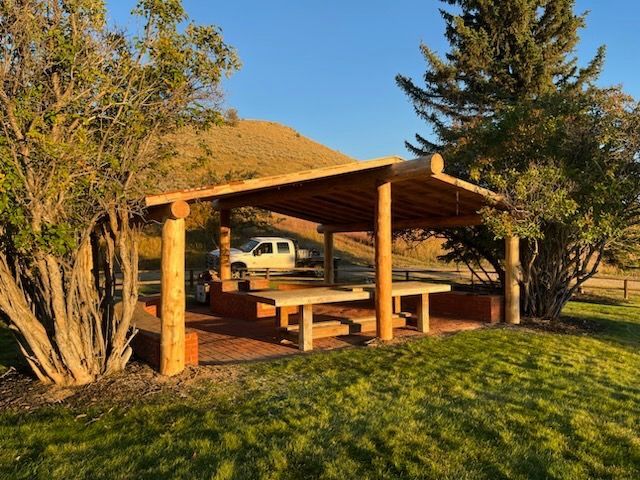 A white truck is parked under a wooden shelter in the grass.