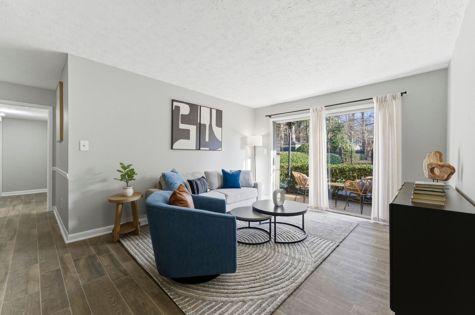 Bright living room with blue chairs, coffee table, wall art, and sliding glass doors to a patio garden.