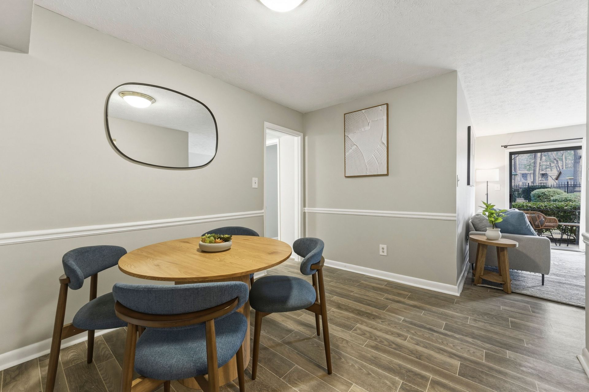 Dining nook with round wood table, blue chairs, wall mirror, and bright open living area beyond.