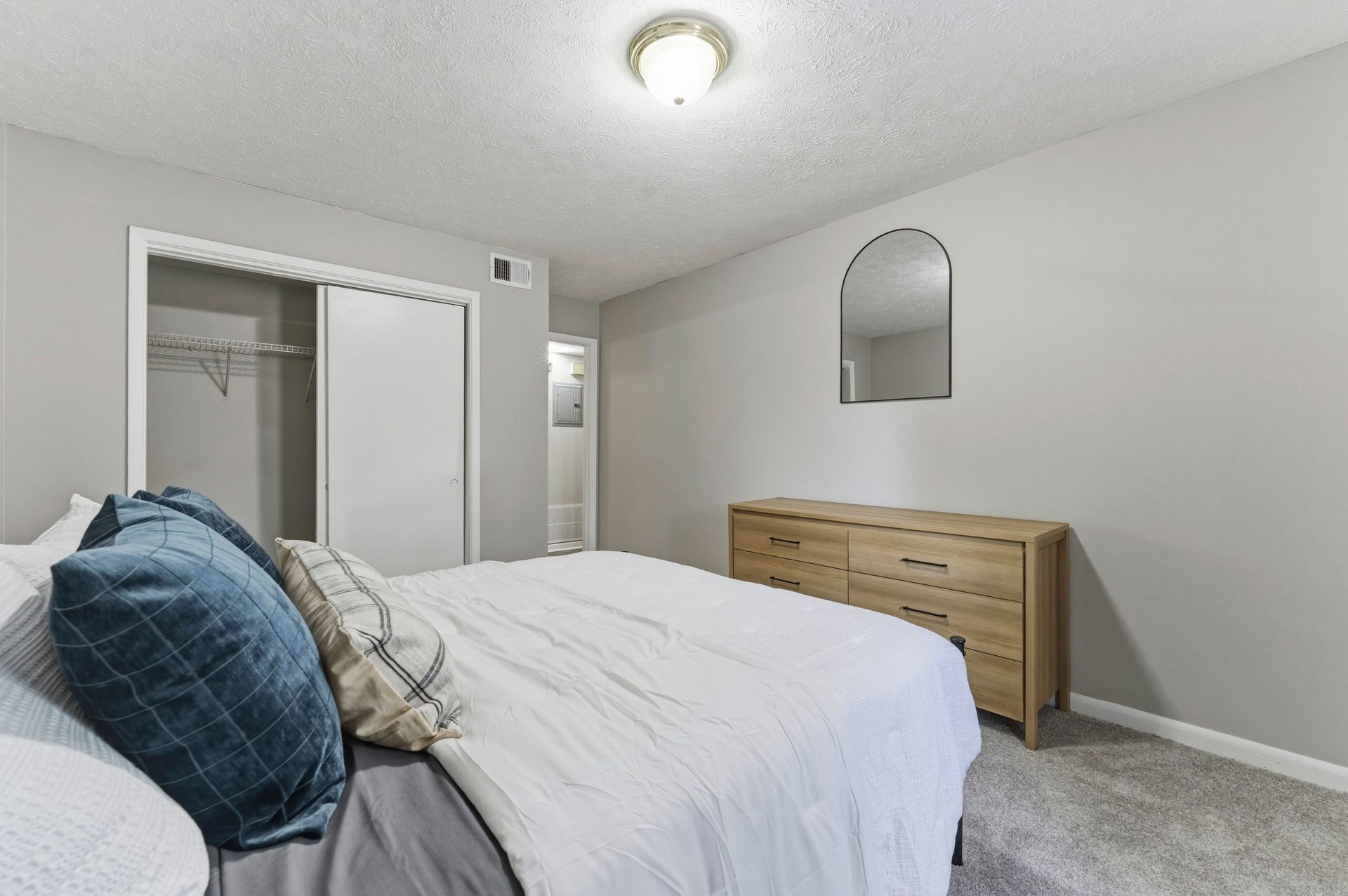 Bright bedroom with white bed, blue pillows, dresser, mirrored wall decor, and open closet door.