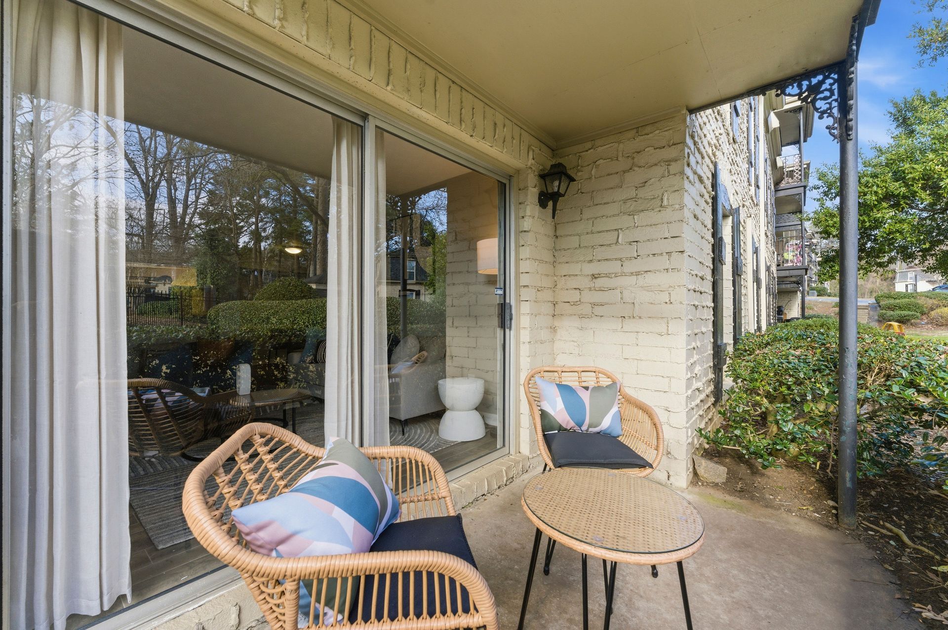 Covered patio with wicker chairs and small table beside sliding glass doors and a brick wall.
