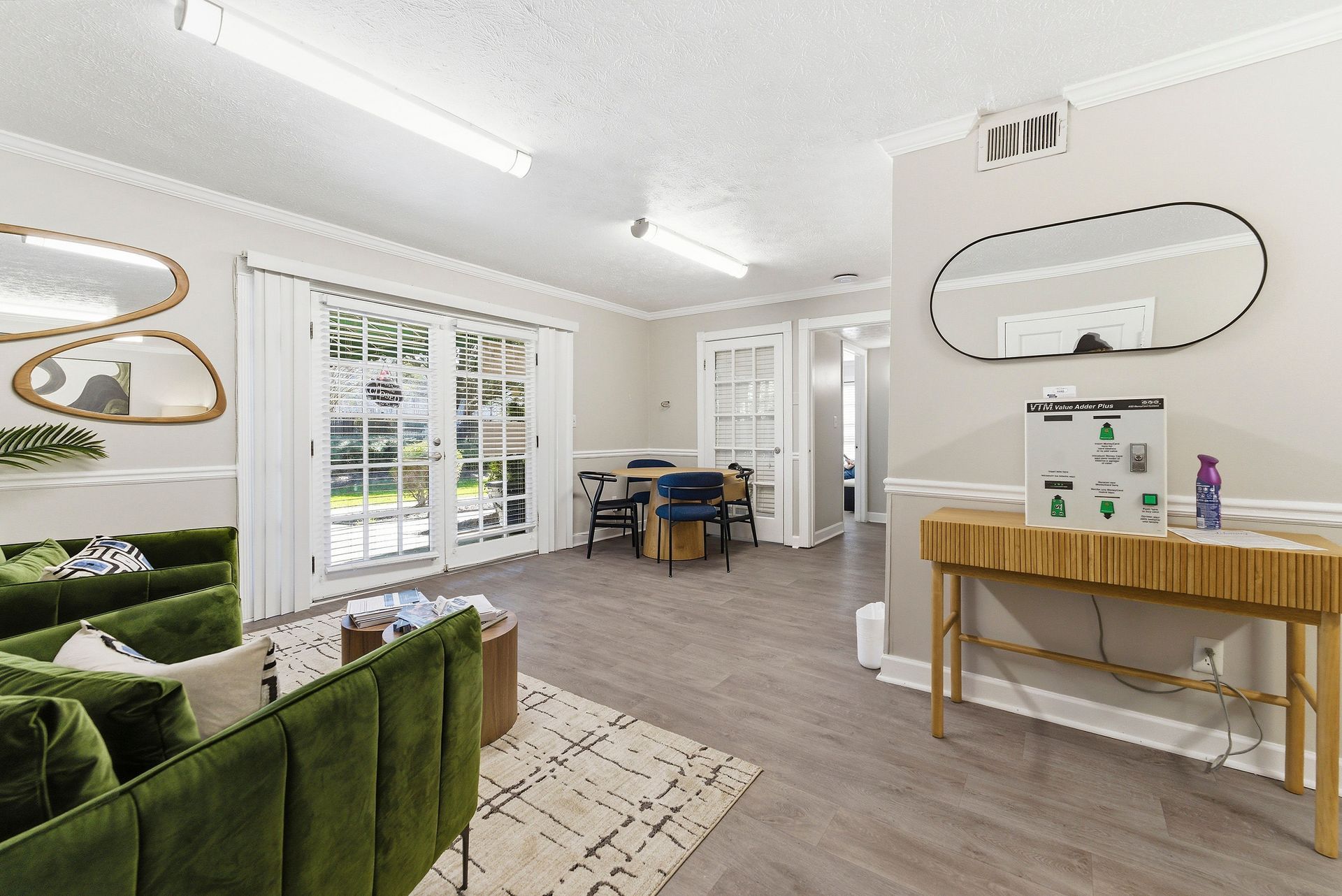 Bright living room with green sofa, white walls, desk, mirror, and large windows with garden view