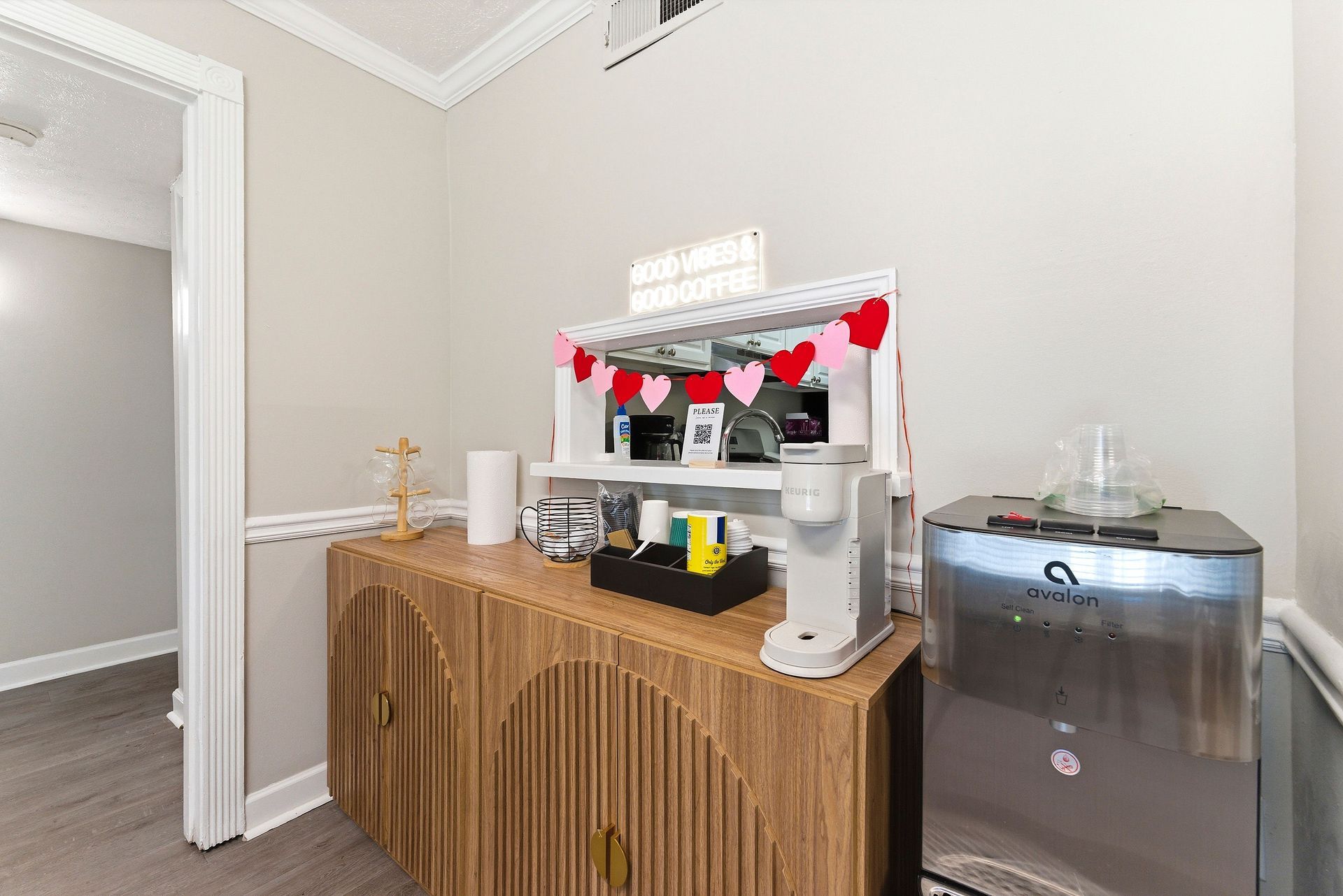 Buffet table with coffee machine, cups, and red heart garland beside a water cooler in a hallway