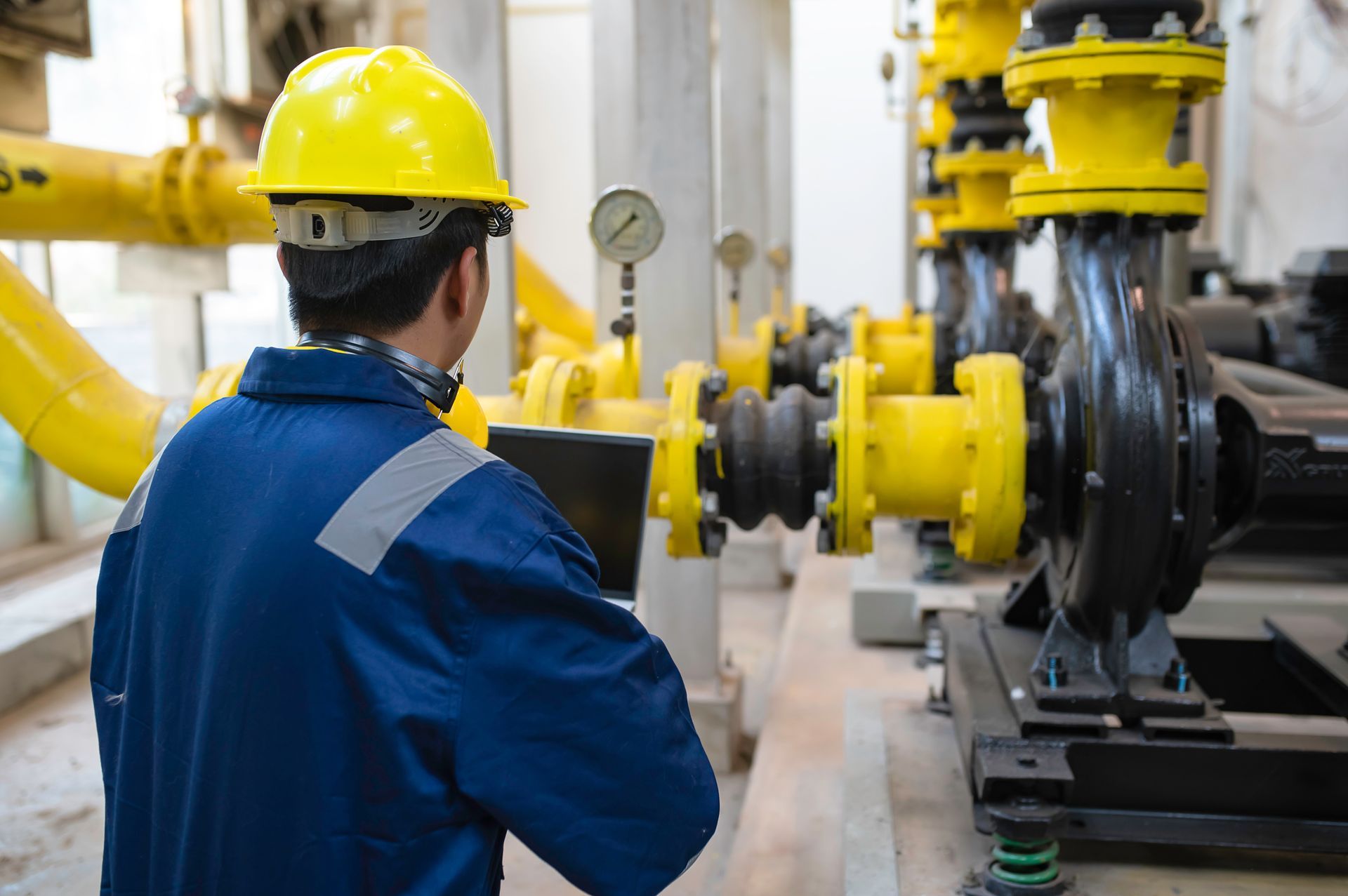Worker in blue coveralls and yellow hard hat inspecting industrial piping and machinery in a plant Worker in blue coveralls and yellow hard hat inspecting industrial piping and machinery in a plant